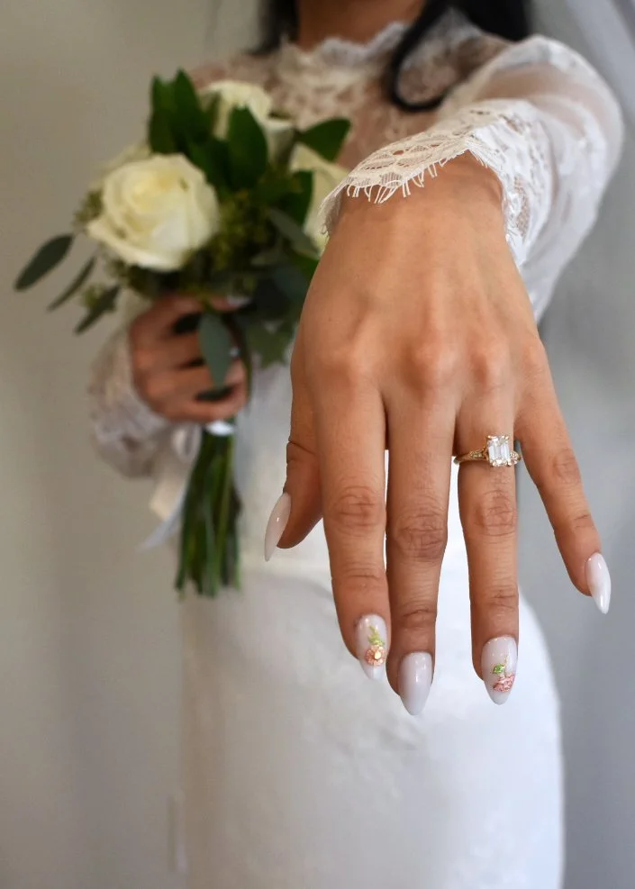 Woman wearing a lace white dress showing her hand with a large engagement ring, holding a bouquet of white roses and greenery.