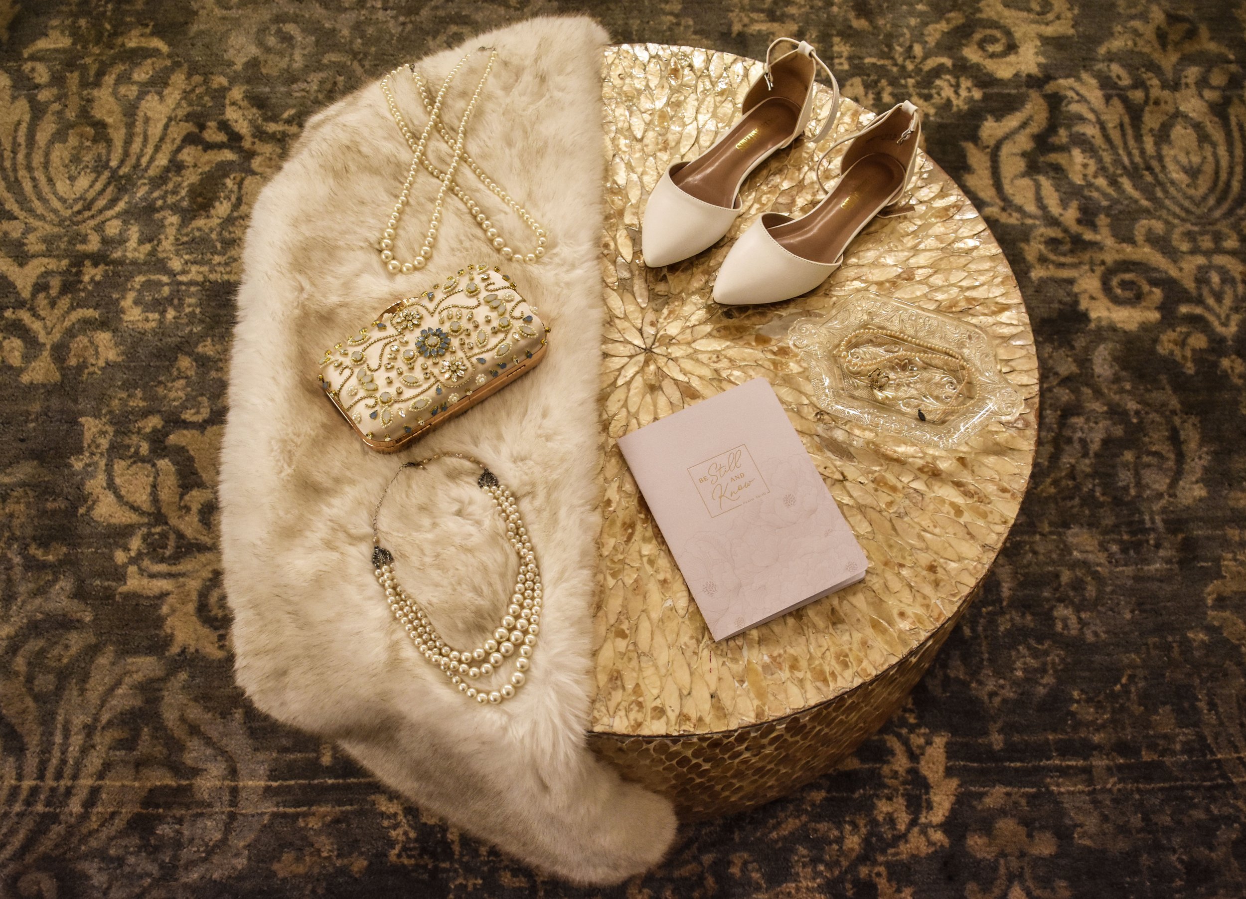 A round table with a textured, gold surface displays various accessories and items, including a white fur stole, pearl necklaces, a decorative clutch, white high-heeled shoes, a glass dish with jewelry, and a pink booklet.