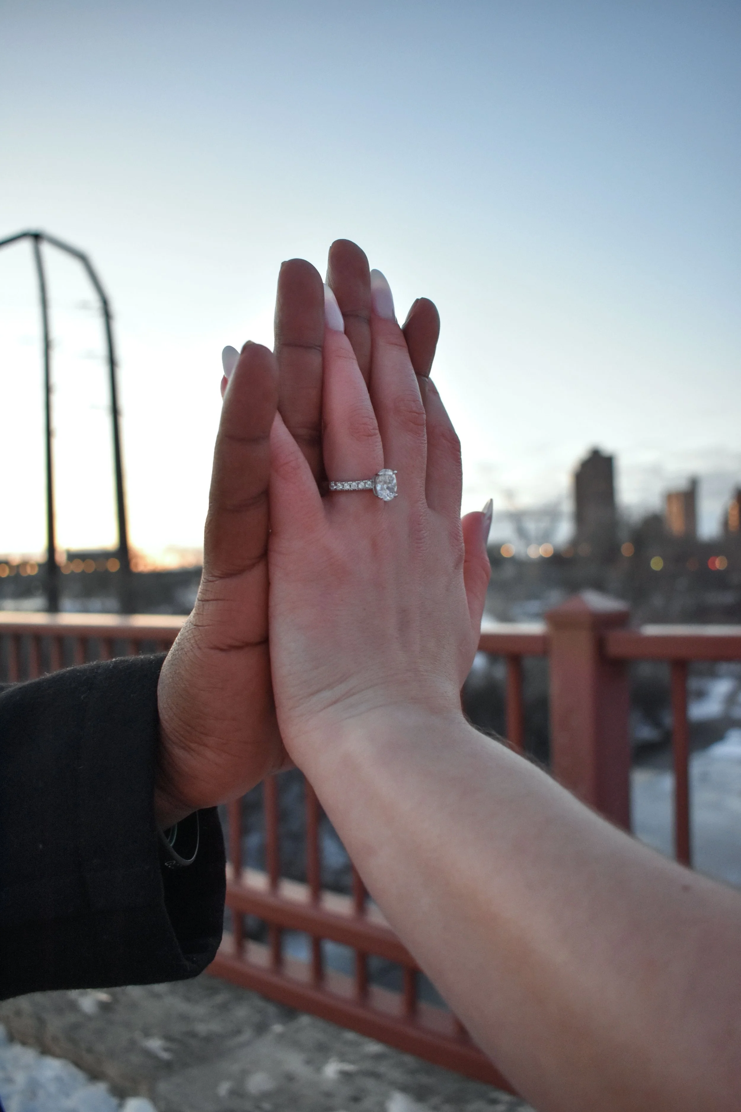 Two hands, one with an engagement ring, pressed together against an outdoor backdrop with a railing and city skyline at sunset.