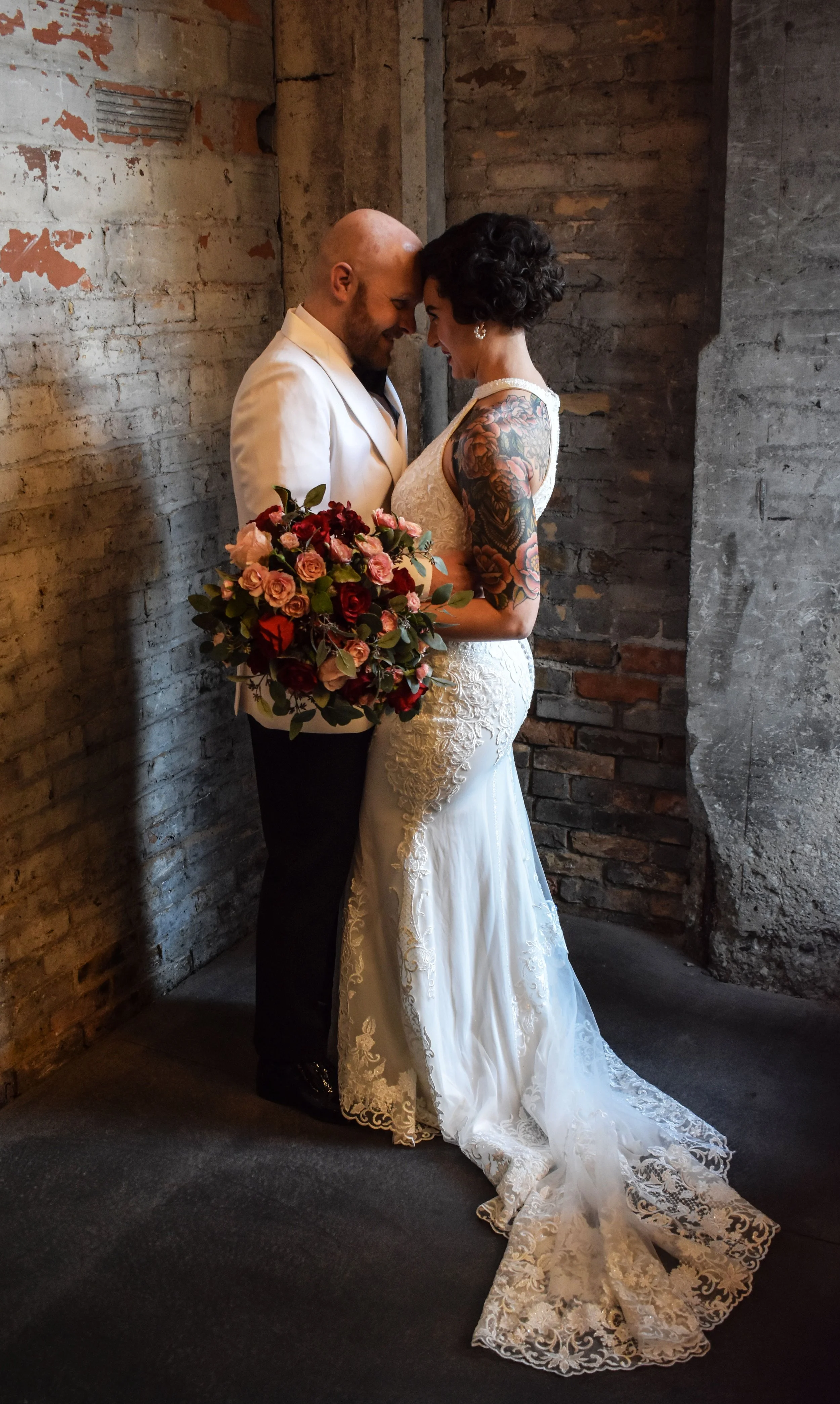 A bride and groom in a close embrace, dressed in wedding attire, with the bride holding a bouquet of roses, standing against a rustic brick wall.