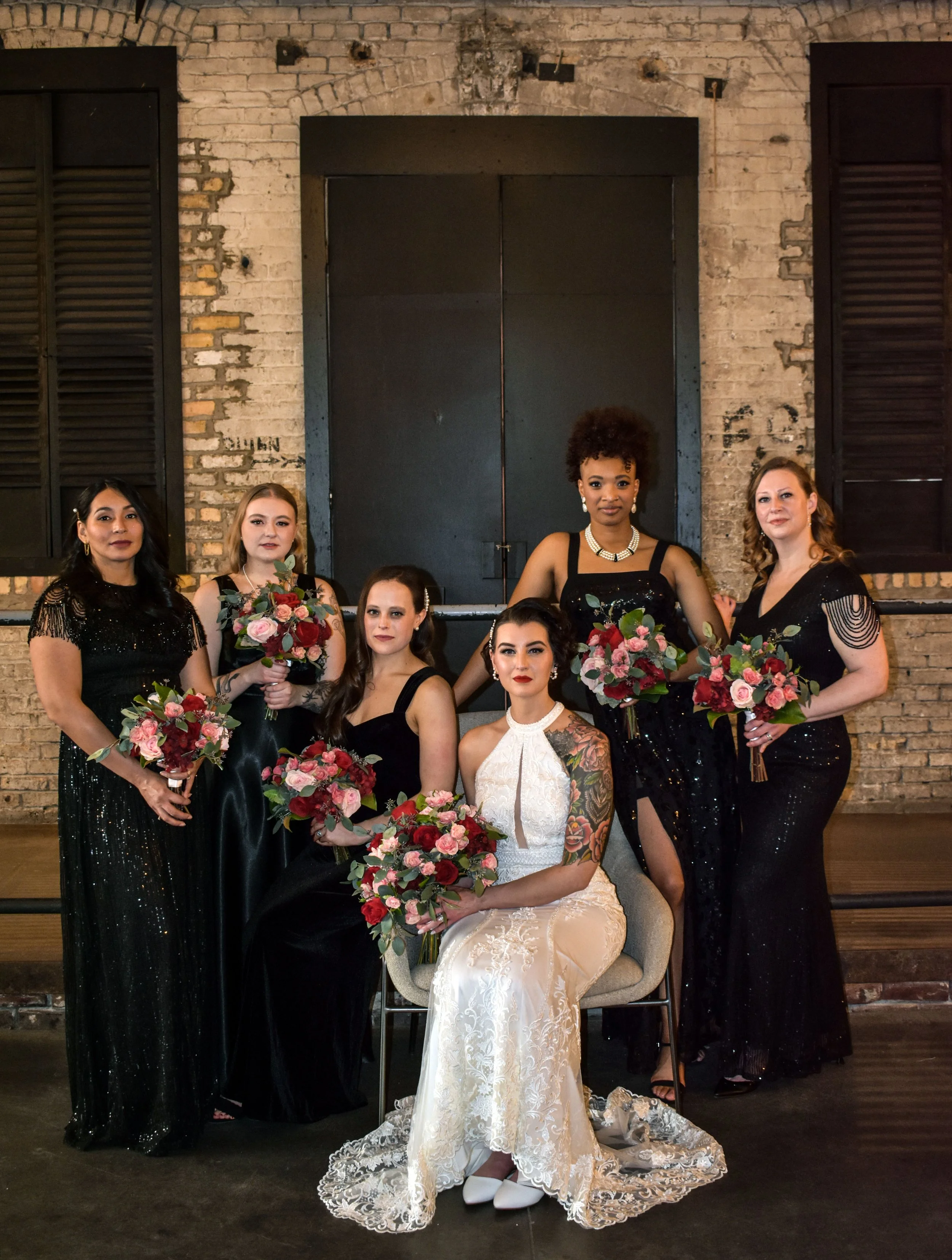 Group of seven women dressed in elegant black and white dresses, holding bouquets of pink and red flowers, posed against an exposed brick wall with black shutters and a black door.