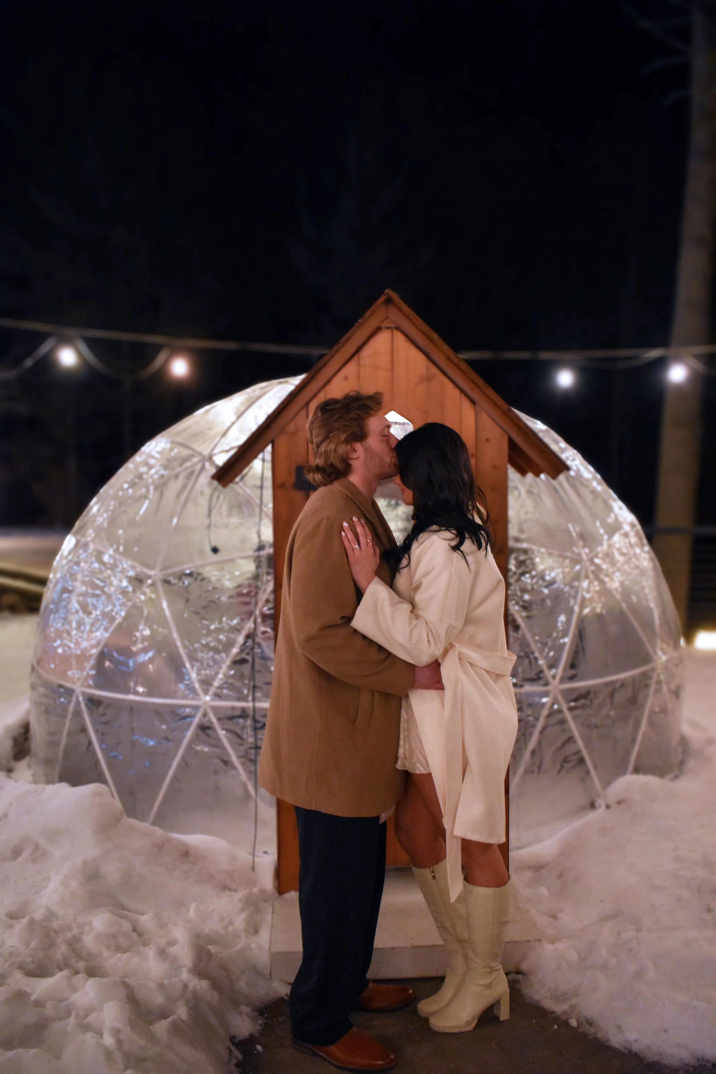 A couple kissing in front of a small wooden house with a clear geodesic dome behind it, surrounded by snow at night, with string lights above.