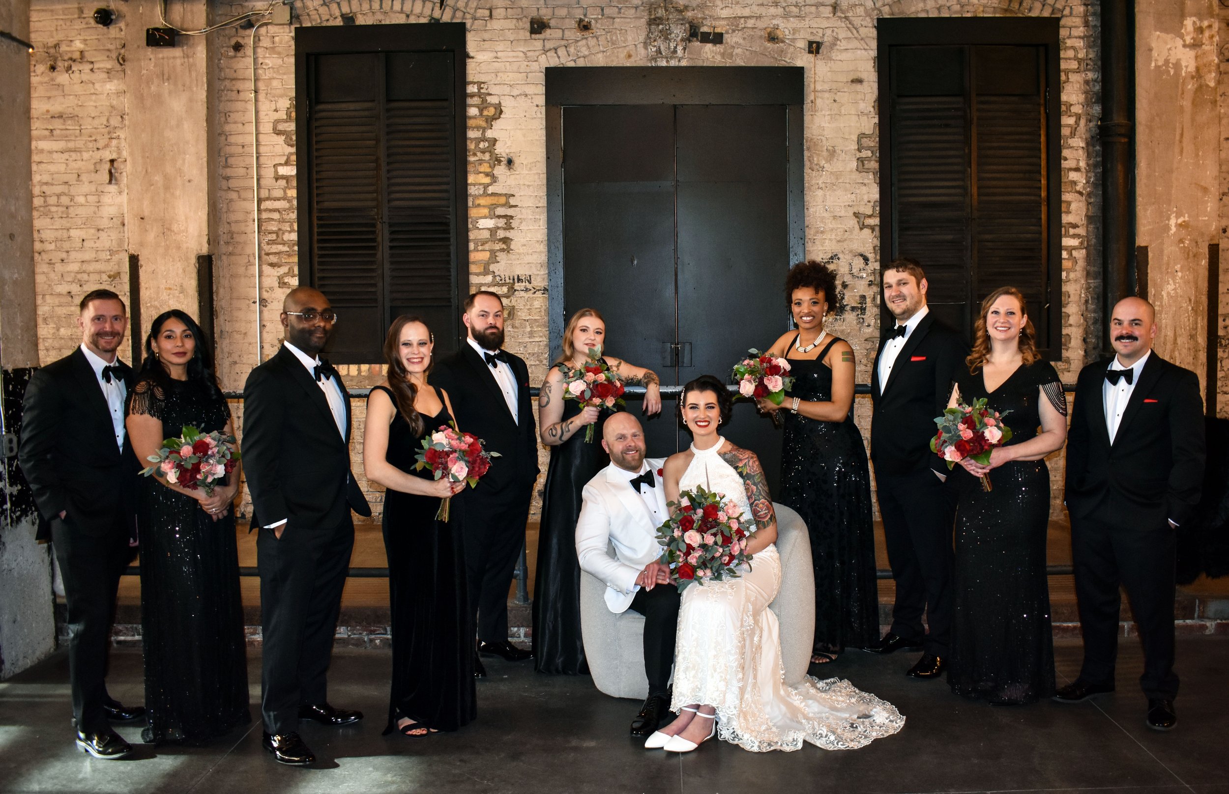 Wedding party consisting of eleven people posing indoors against a brick wall with black shutters. Central couple seated, bride in white lace gown and groom in white tuxedo, holding large bouquets of pink and red roses. Surrounding wedding party memb