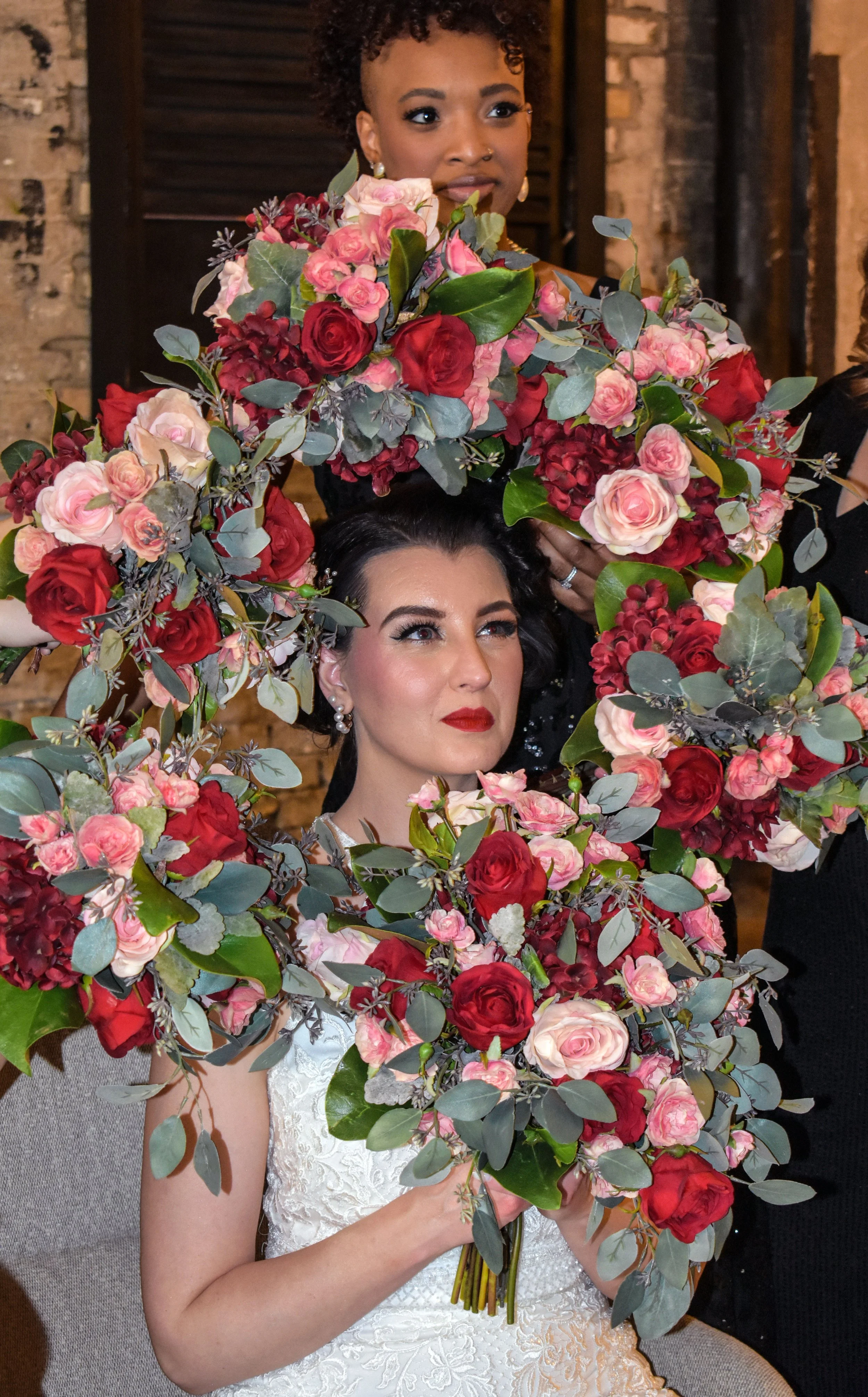 A bride with dark hair and red lipstick holding a large bouquet of pink, red, and burgundy flowers with green leaves. Behind her, a woman with curly hair and earrings is placing flowers, with a brick wall in the background.