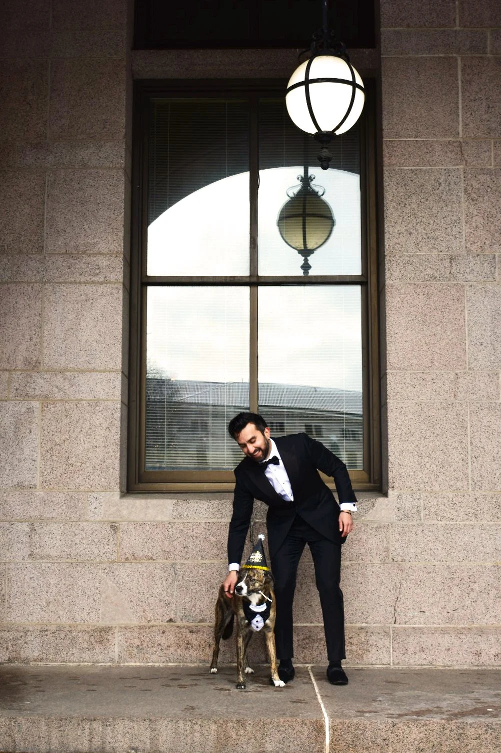 A man in a tuxedo smiling while bending down to pet a dog dressed in a birthday hat and bow tie, standing in front of a brick wall and window with reflections of lanterns.