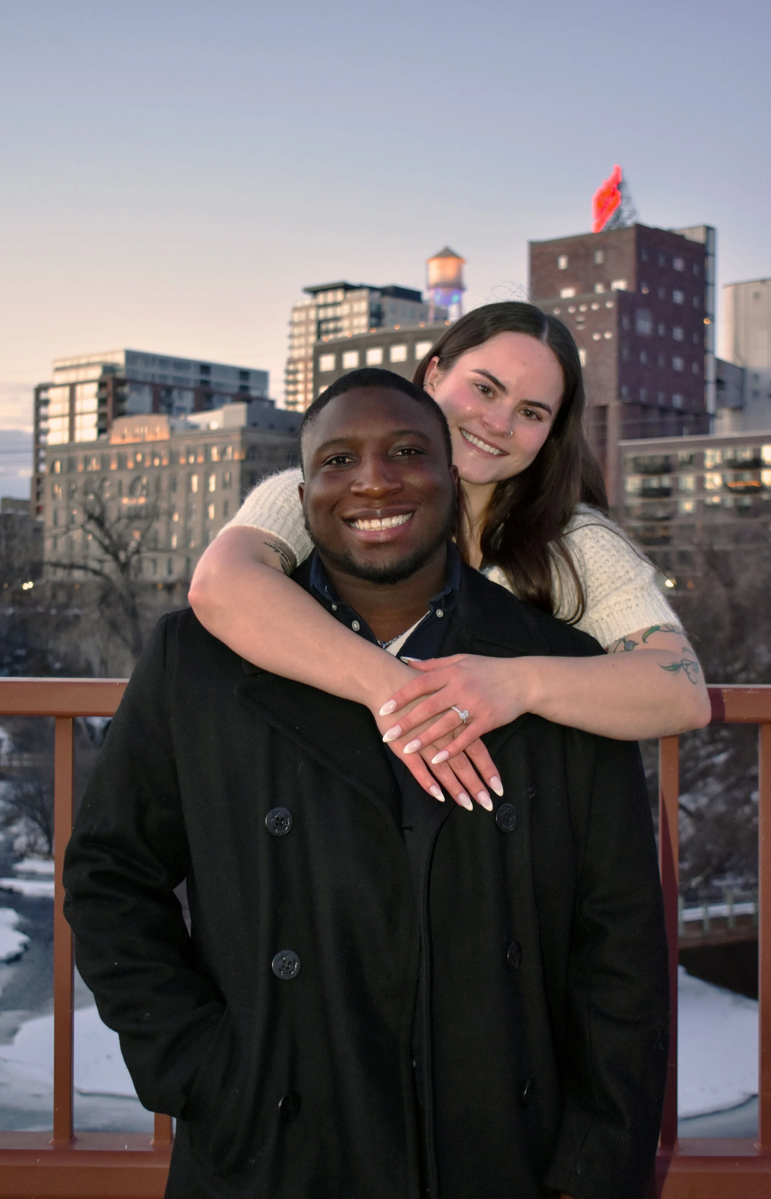 A smiling man with black hair and a clean-shaven face wearing a black coat, with a woman with long dark hair and a nose piercing, smiling, hugging him from behind. She has tattoos on her forearm and is wearing a white sweater, set against a city skyl