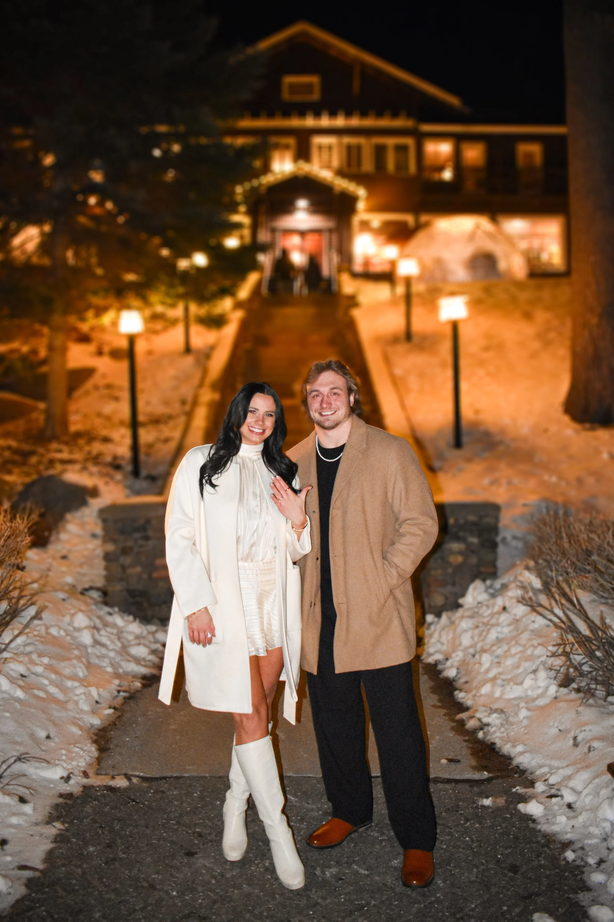 A couple standing outdoors on a snowy path at night, dressed warmly, with a large house illuminated in the background.