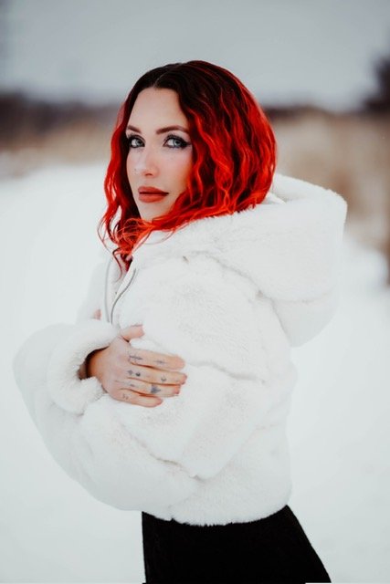 Woman with curly red hair in a white, fluffy jacket standing outdoors in a snowy landscape.