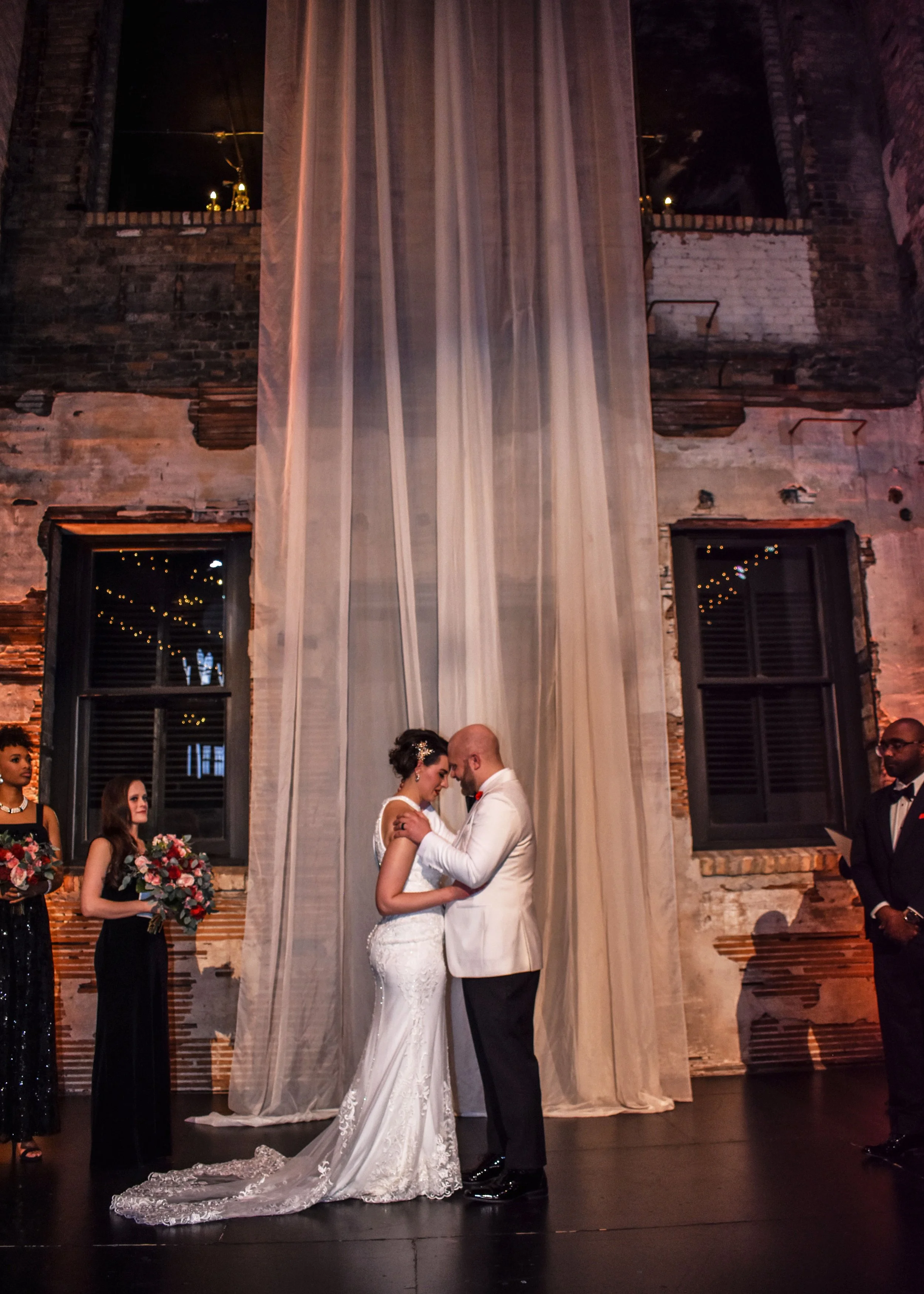 A bride and groom share a wedding kiss in front of a large translucent curtain, with bridesmaids holding bouquets and a groomsman standing nearby in a dimly lit, industrial-style venue with exposed brick walls and windows.