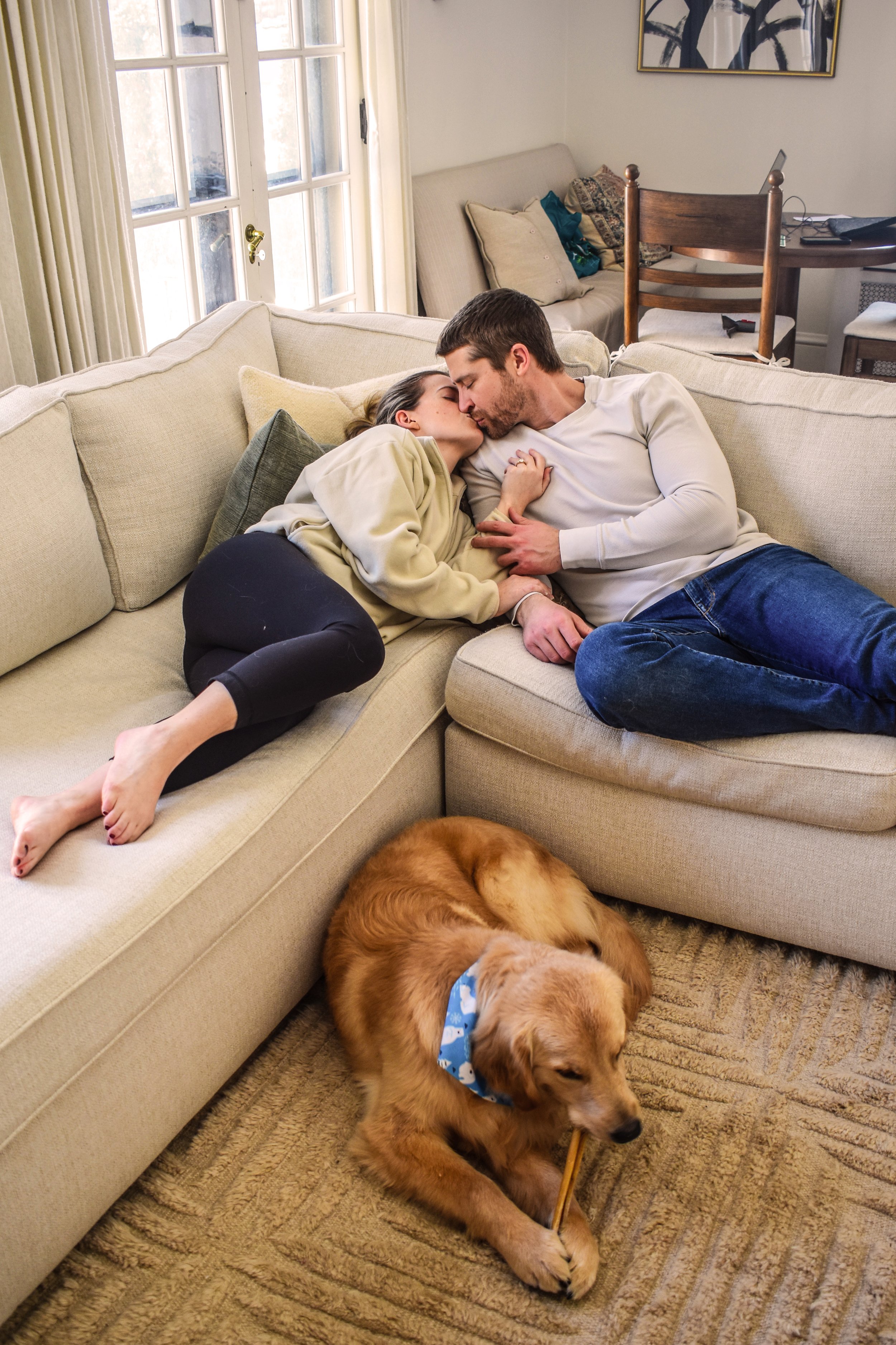 A couple is lying on a beige sectional sofa, sharing a kiss, with a golden retriever puppy chewing a toy on the carpet nearby.