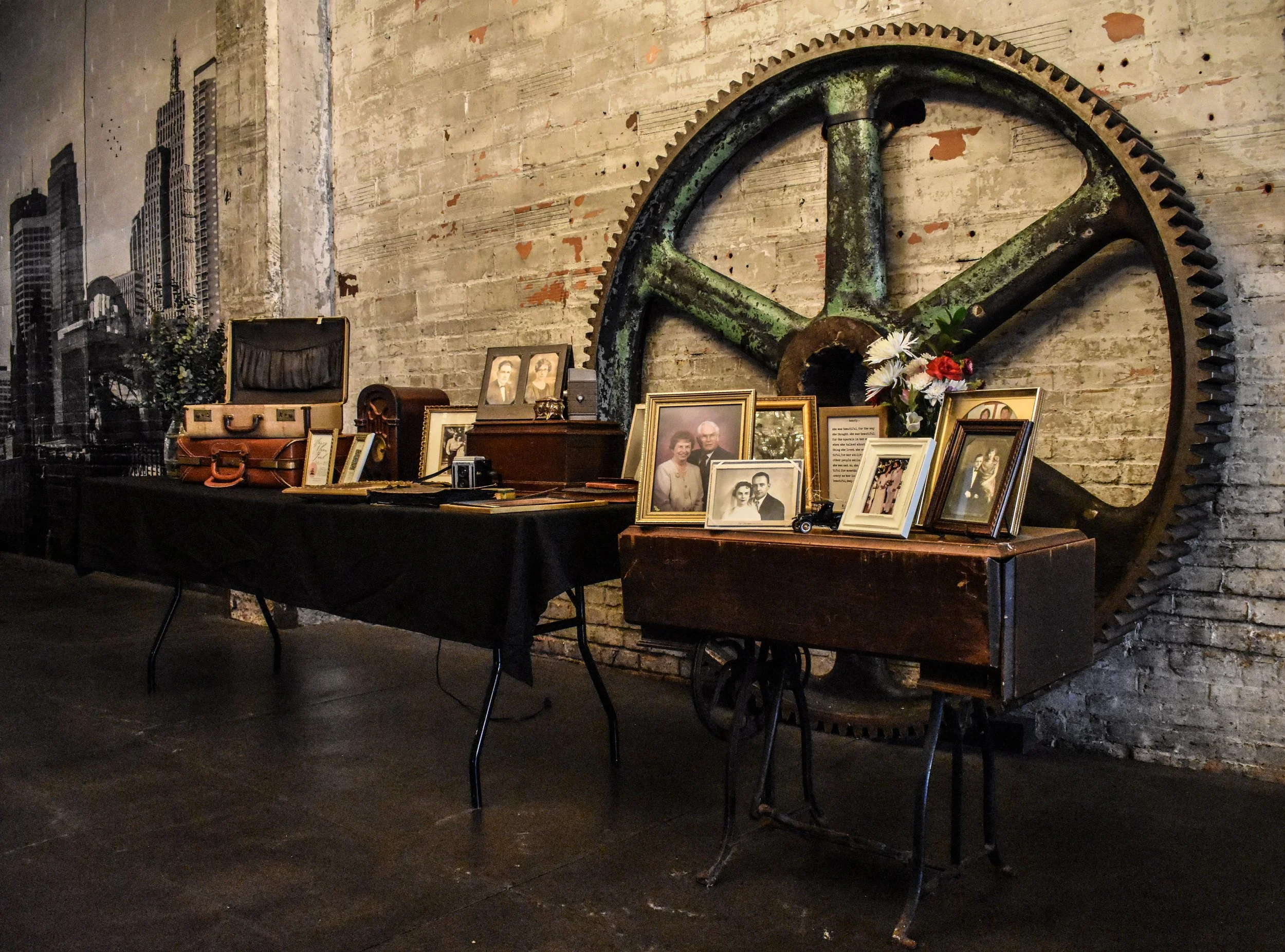 Display of framed photographs, flowers, and vintage suitcases on tables in front of a large gear on an exposed brick wall, with some cityscape artwork on the wall to the left.