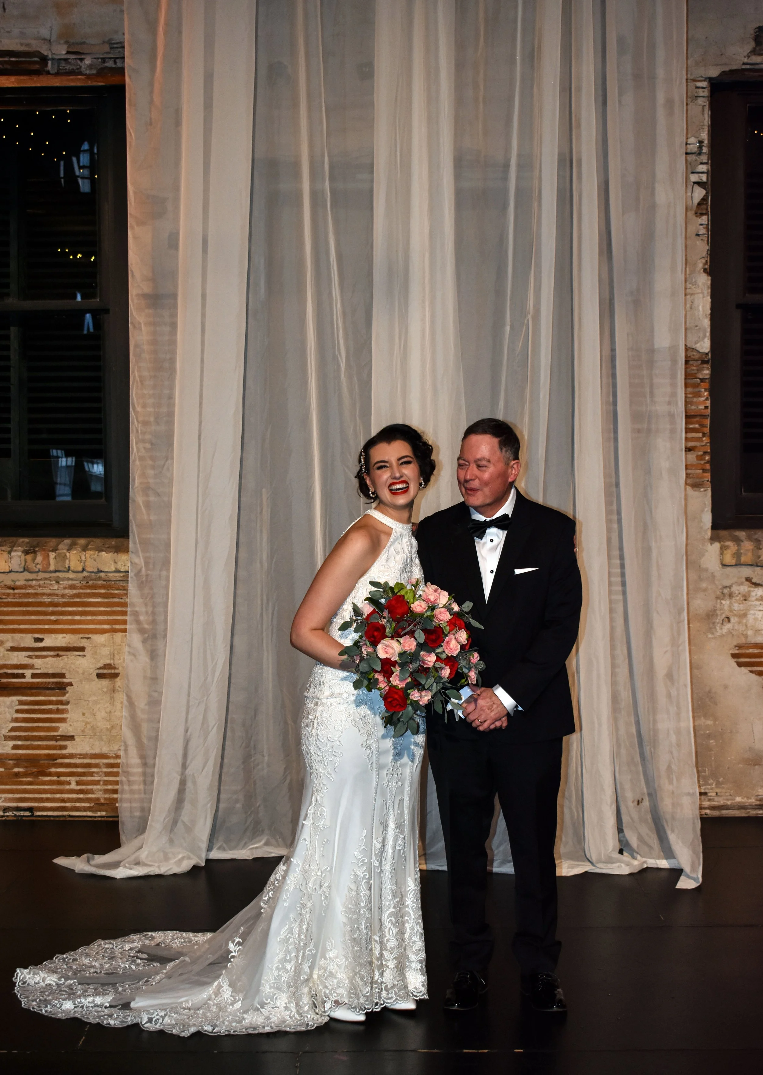 A bride and groom standing together during their wedding celebration, with the bride holding a large bouquet of pink and red roses, smiling happily in front of a curtain backdrop.