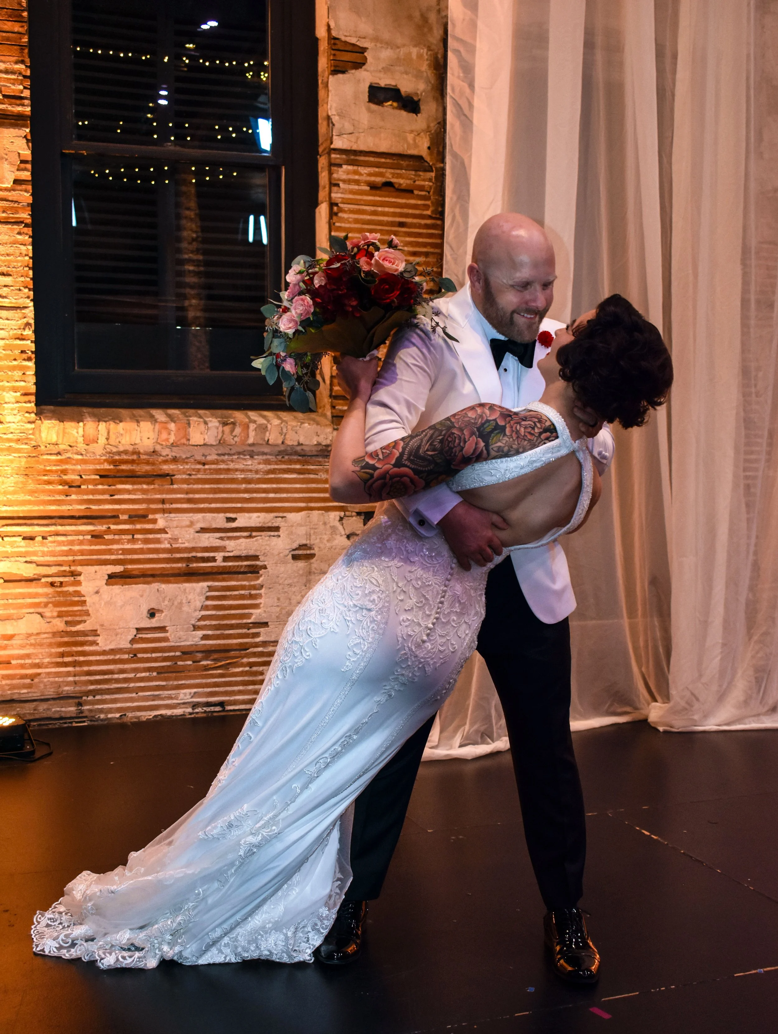 A wedding couple dancing, with the groom holding a bouquet and the bride wearing a lace wedding dress, in an industrial-style venue with brick walls and curtains.