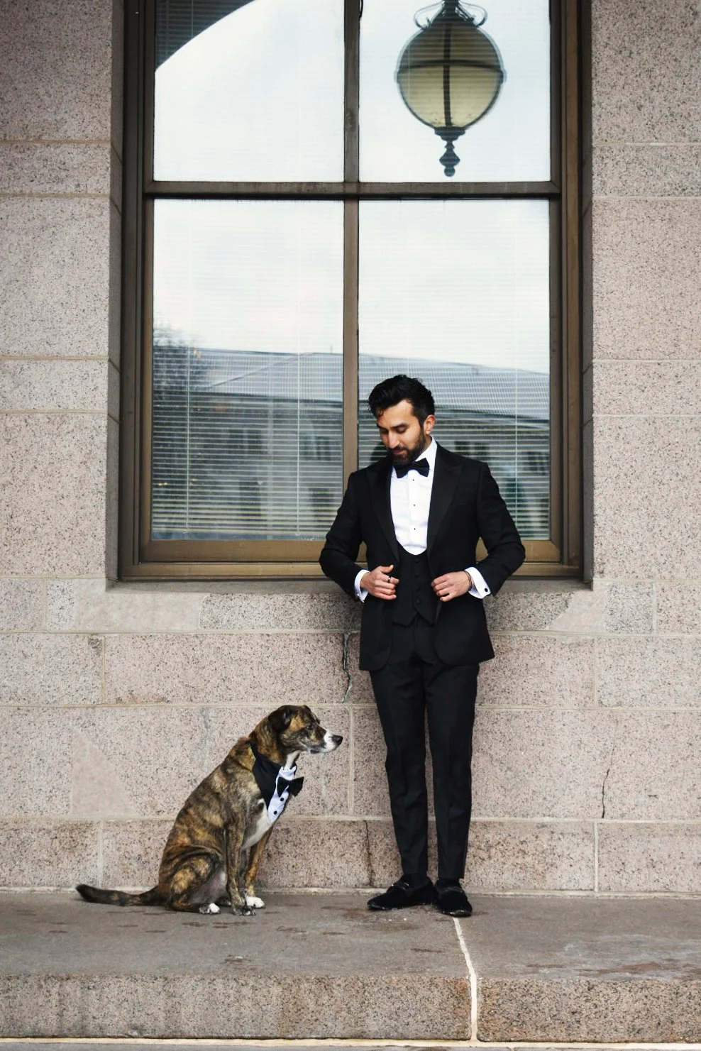Man in a tuxedo standing next to a dog in a tuxedo bow tie outside a building with a large window and hanging lamp.