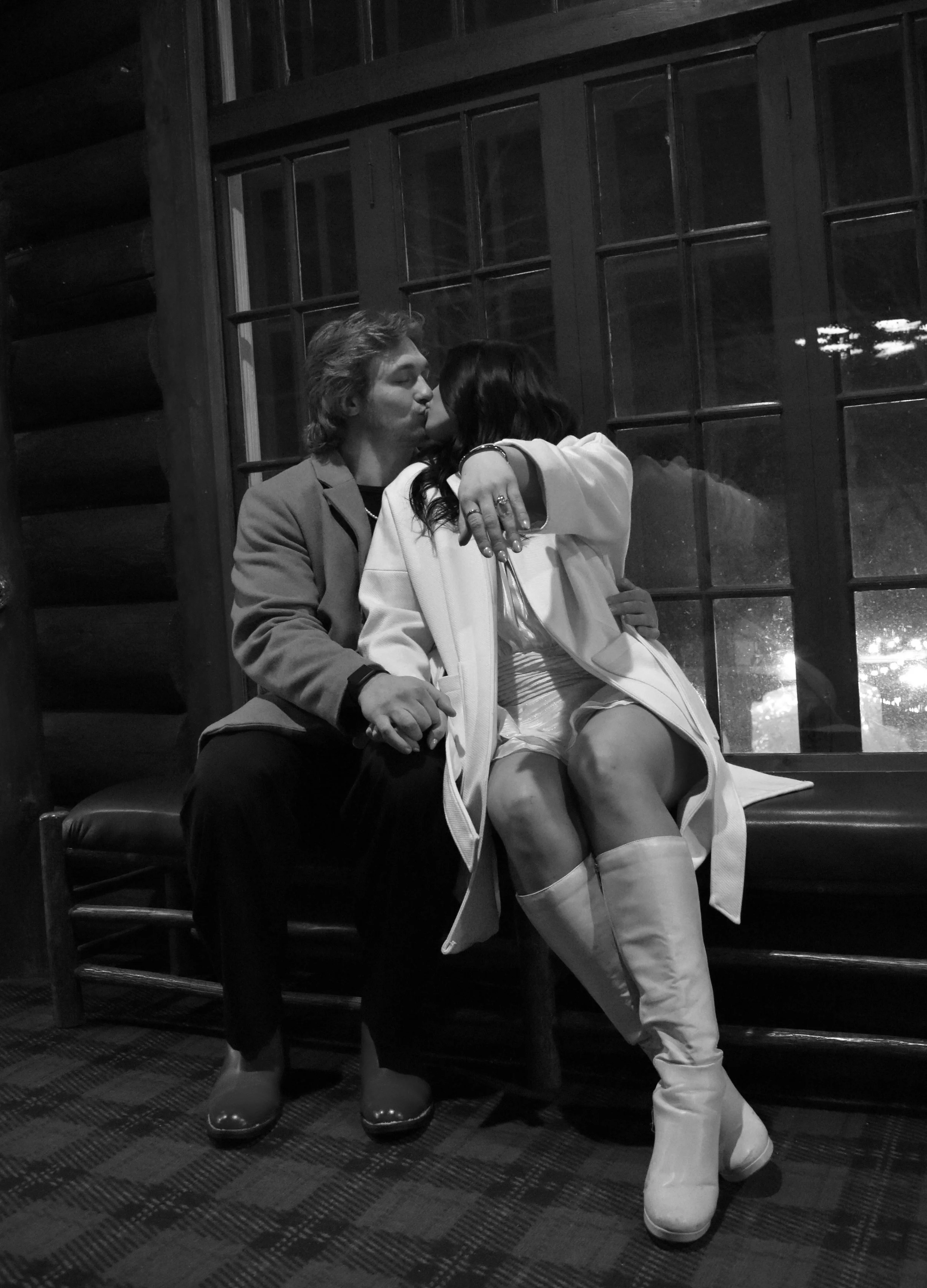 A young man and woman sharing a kiss, sitting on a bench in front of a window inside a rustic wooden room at night.