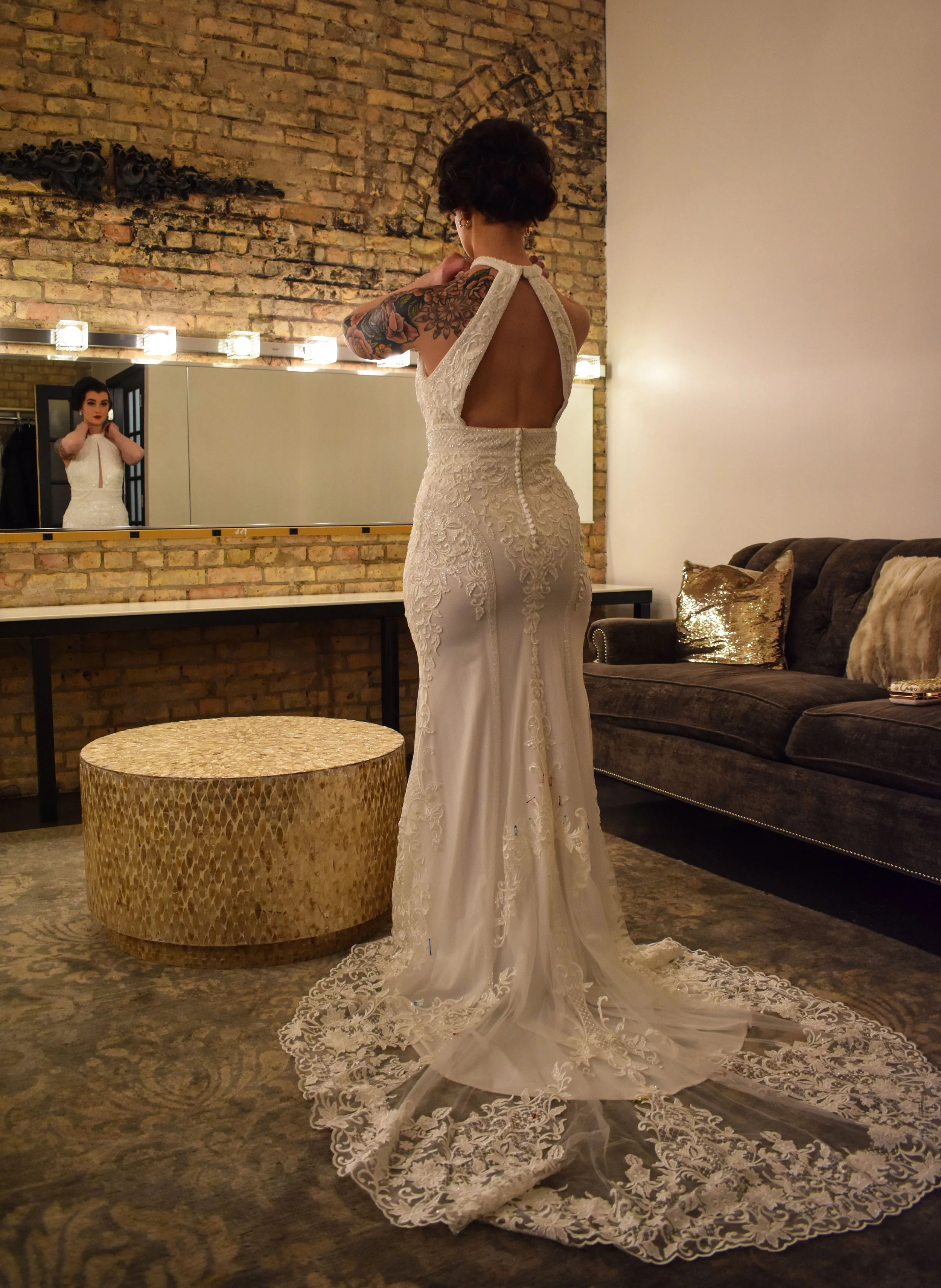 A woman trying on a white wedding dress in a bridal fitting room, seen from behind in front of a mirror, with a brick wall and sofa in the background.