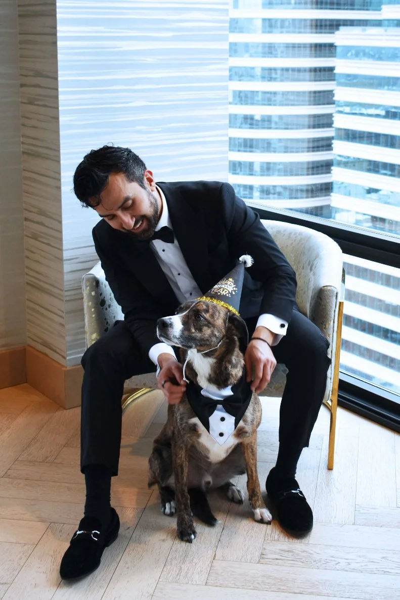 A man in a tuxedo sitting in a chair, smiling and looking at a dog dressed in a tuxedo costume with a hat, inside a modern building with large windows showing a city skyline.
