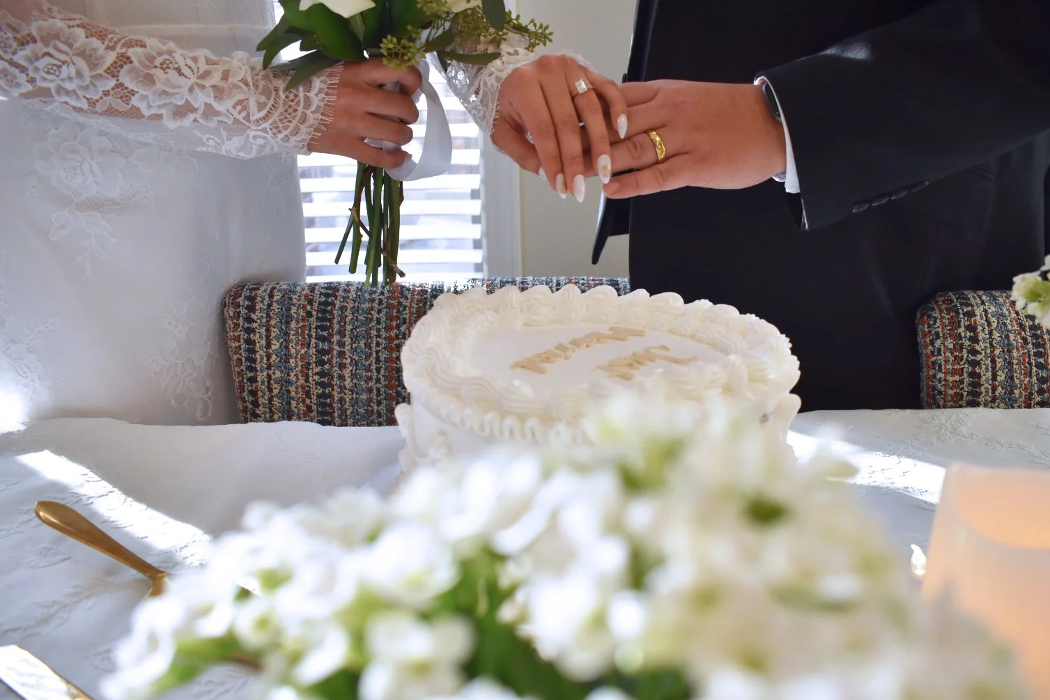 A bride and groom holding hands over a wedding cake on a table, with a bouquet of flowers in the bride's hand and wedding rings visible.