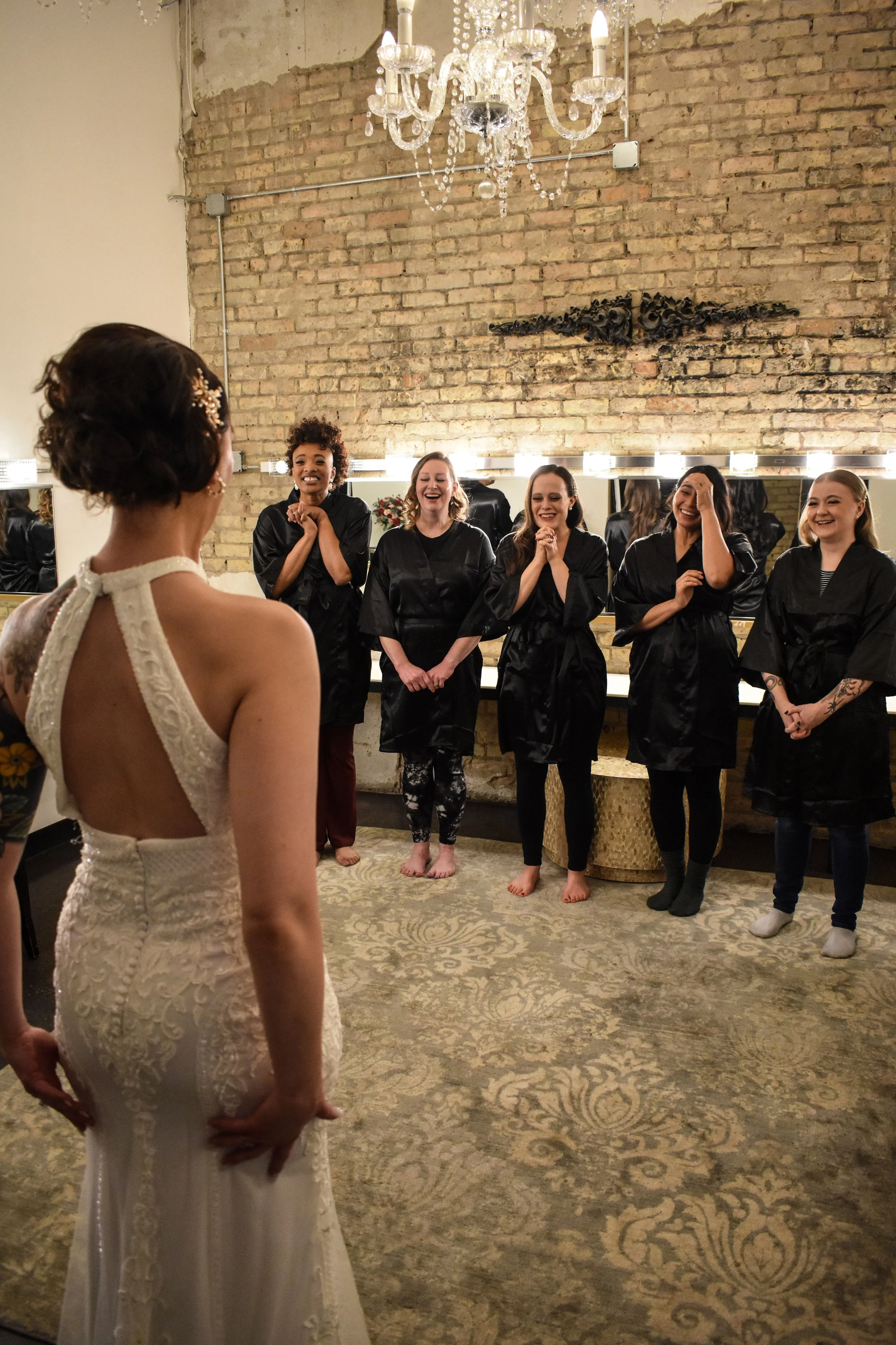 A bride in a white lace wedding gown standing in front of five women dressed in black robes, all smiling and laughing, in a room with a brick wall, a chandelier, and a large mirror.