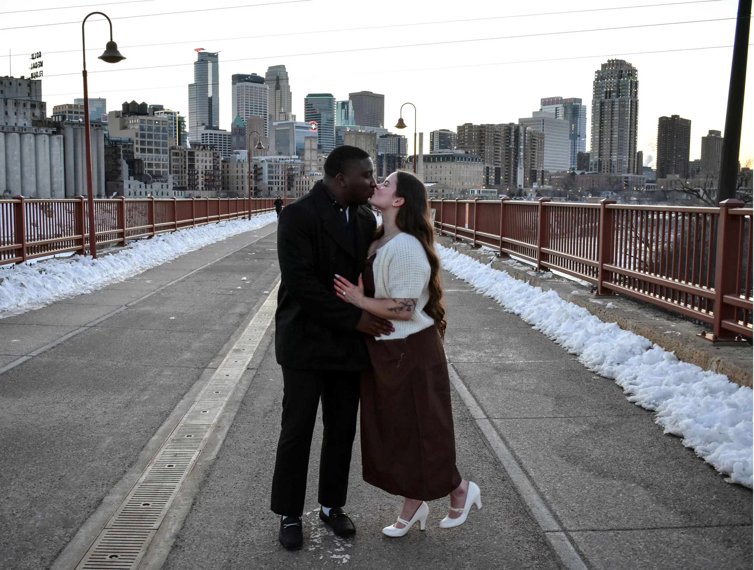 A couple kissing on a city bridge during winter, with snow on the ground and a skyline of tall buildings in the background.