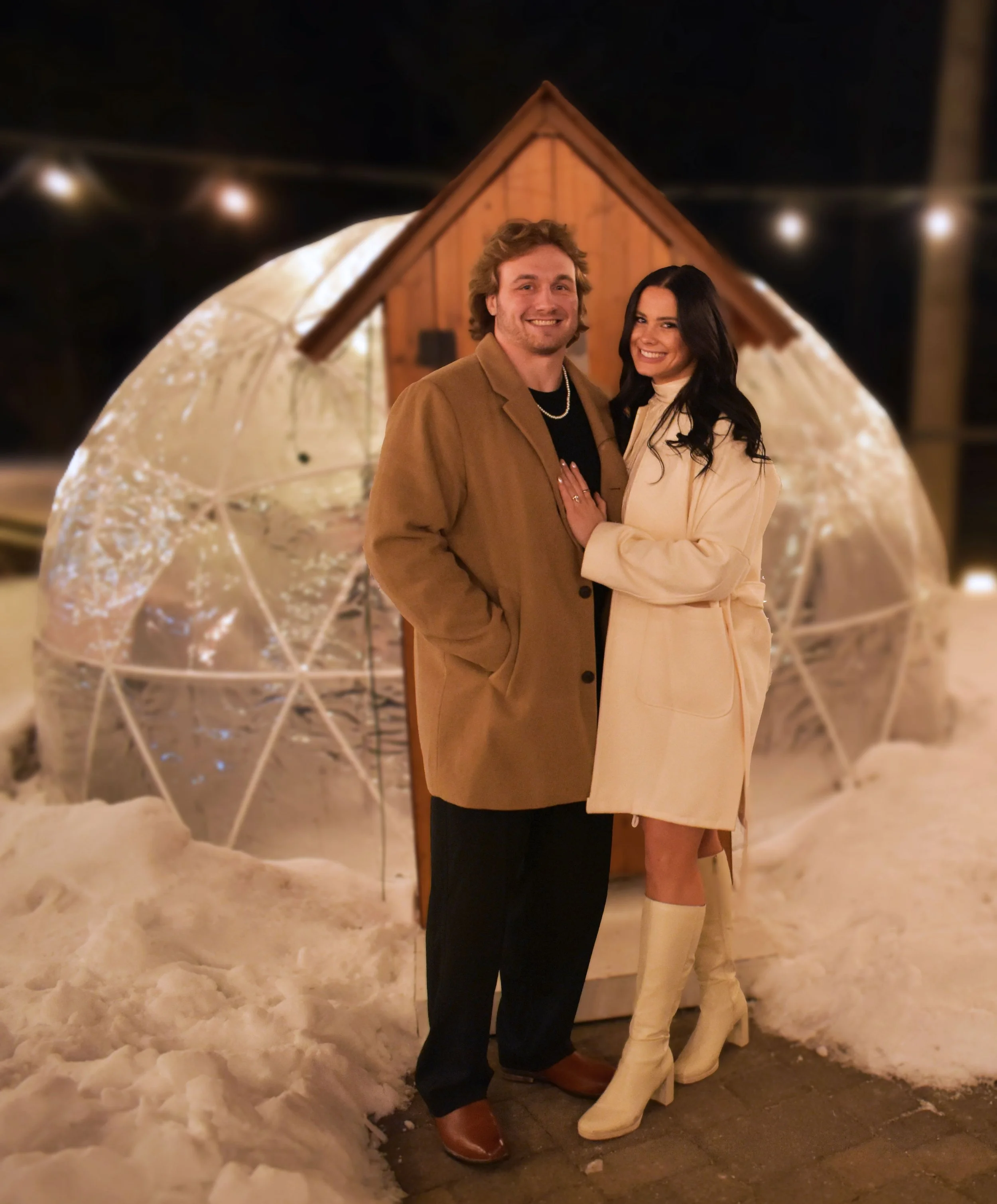 A smiling man and woman standing close together outdoors at night in winter, with snow on the ground, in front of a small wooden shed illuminated by string lights.