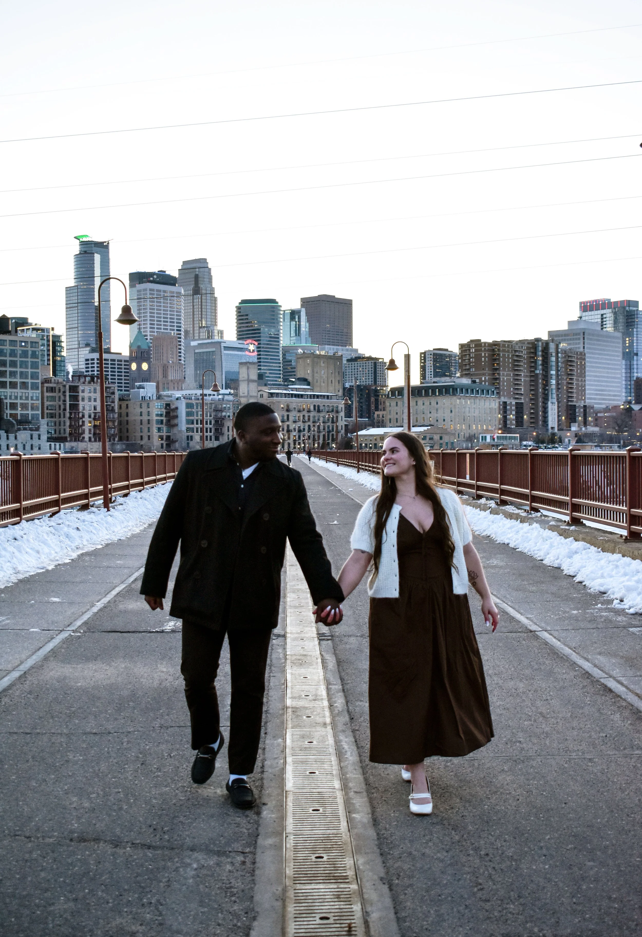 A couple holding hands and walking on a bridge with a city skyline in the background, some snow on the sides of the bridge, and street lamps along the walkway.