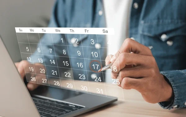 Person holding a stylus and pointing at a transparent digital calendar projecting over a laptop, displaying dates and days of the week.
