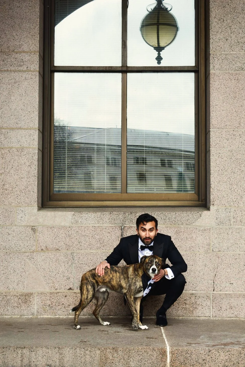 A man in a tuxedo crouching beside a brindle dog on a concrete step in front of a stone wall and large window, with a hanging lamp reflected in the window.