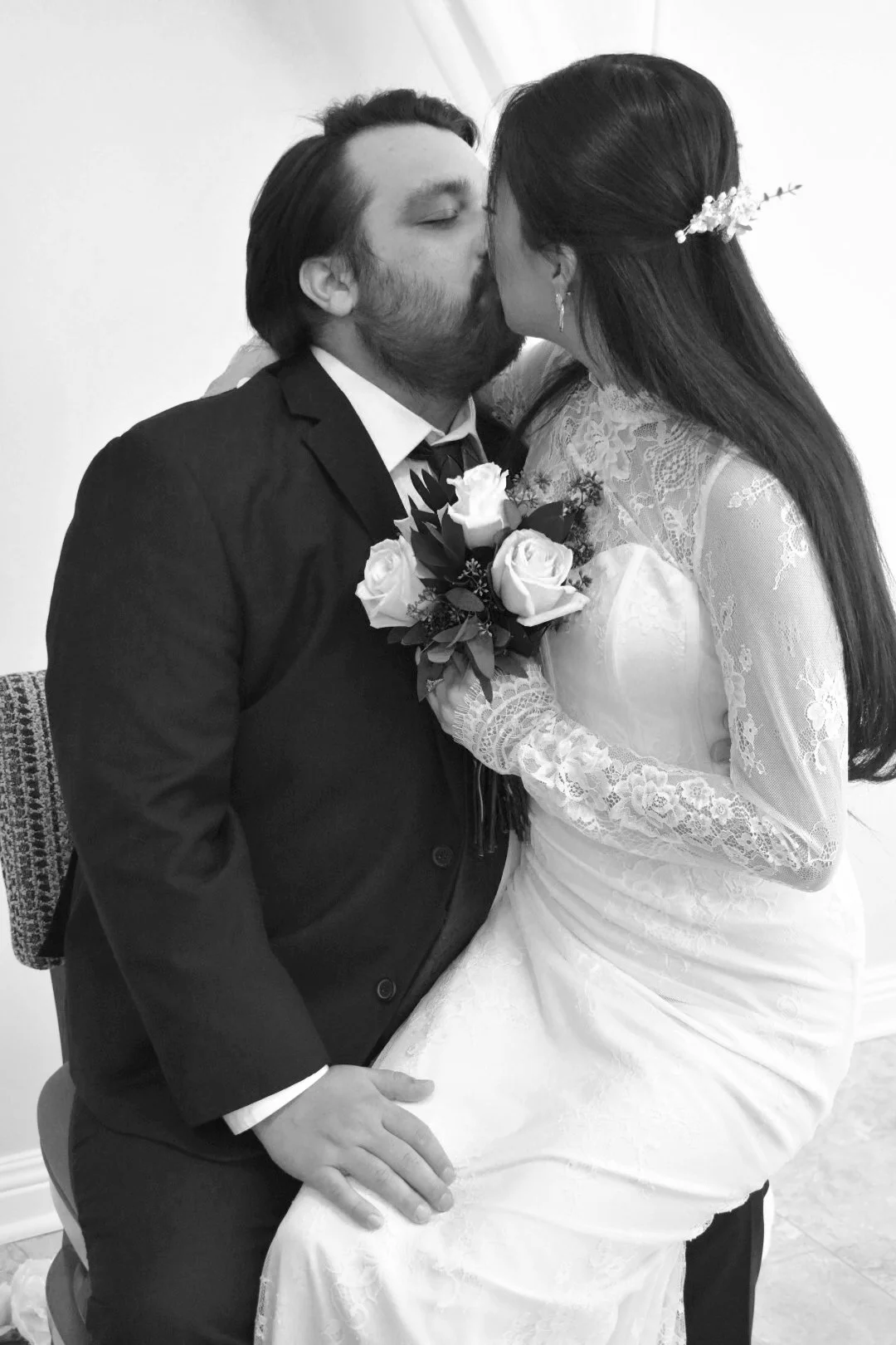 A black-and-white photo of a bride and groom sharing a kiss, with the groom seated and the bride sitting on his lap, holding a bouquet of roses, both dressed in wedding attire.