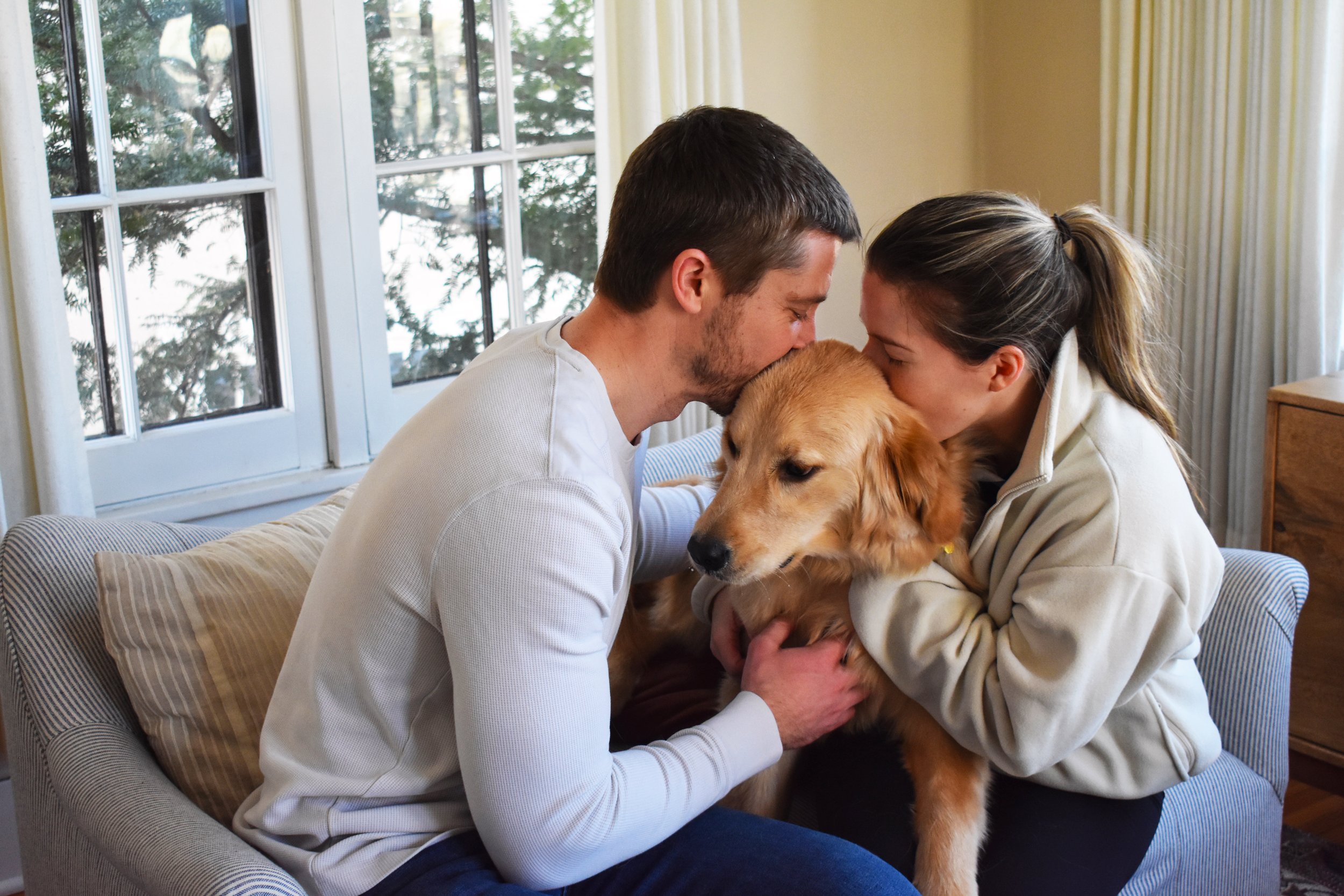 A couple sitting on a couch, kissing their golden retriever dog on the head inside a living room with large windows and natural light.