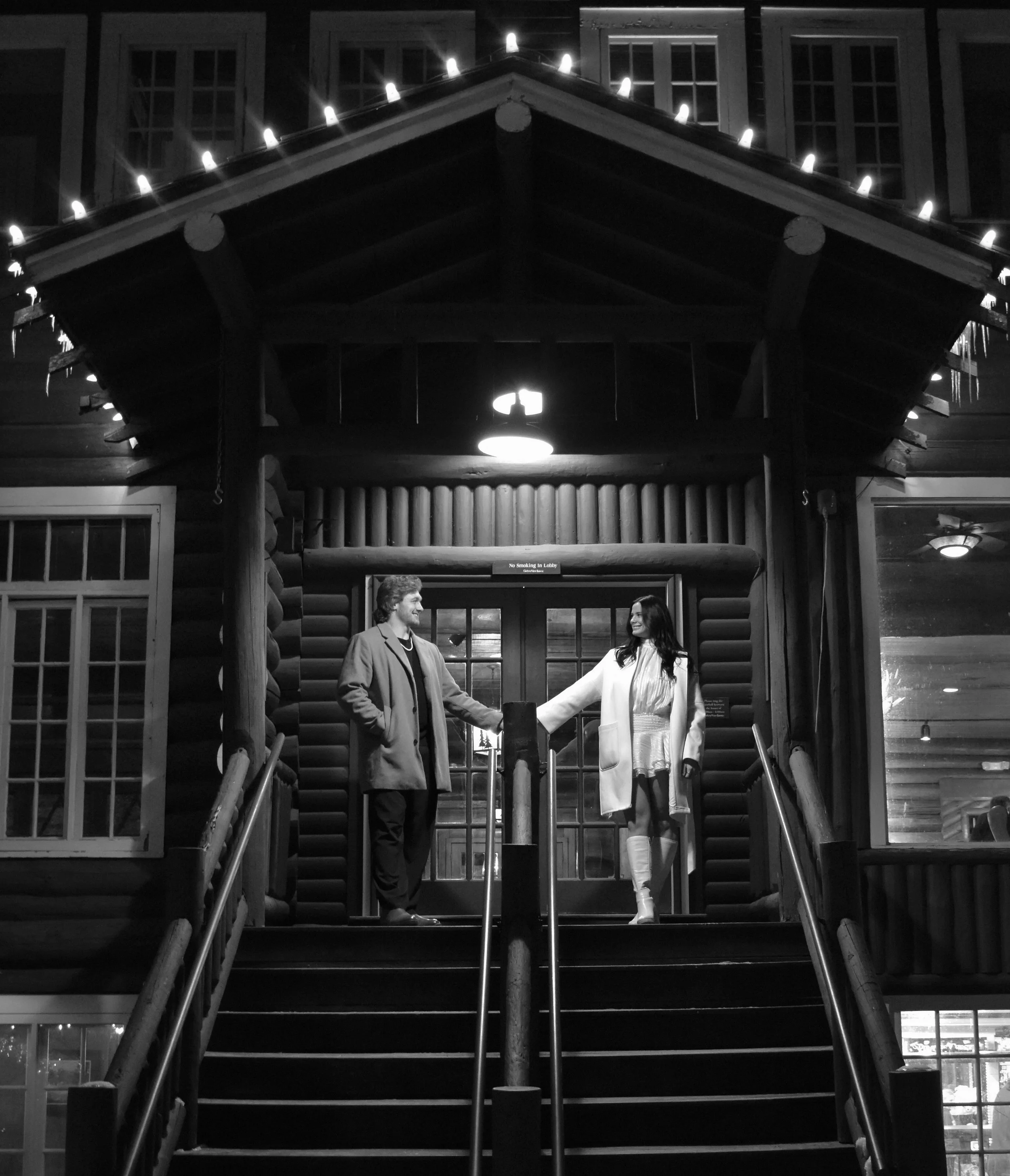 A black and white photo of a man and woman standing on a porch, holding hands, in front of a wooden cabin with string lights overhead, at night.