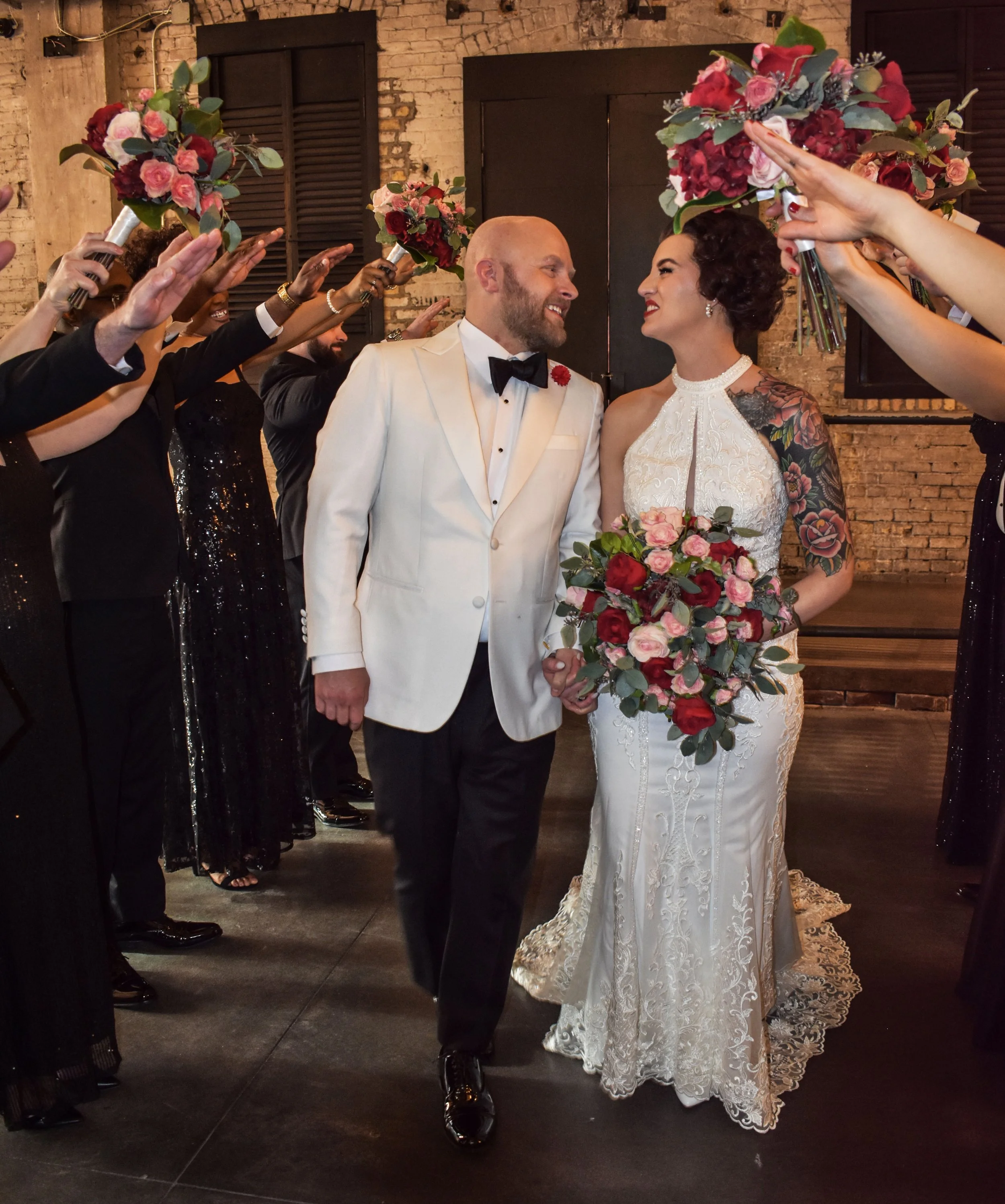 A wedding couple holding hands, walking down the aisle while surrounded by friends and family holding bouquets and raising their arms in celebration.