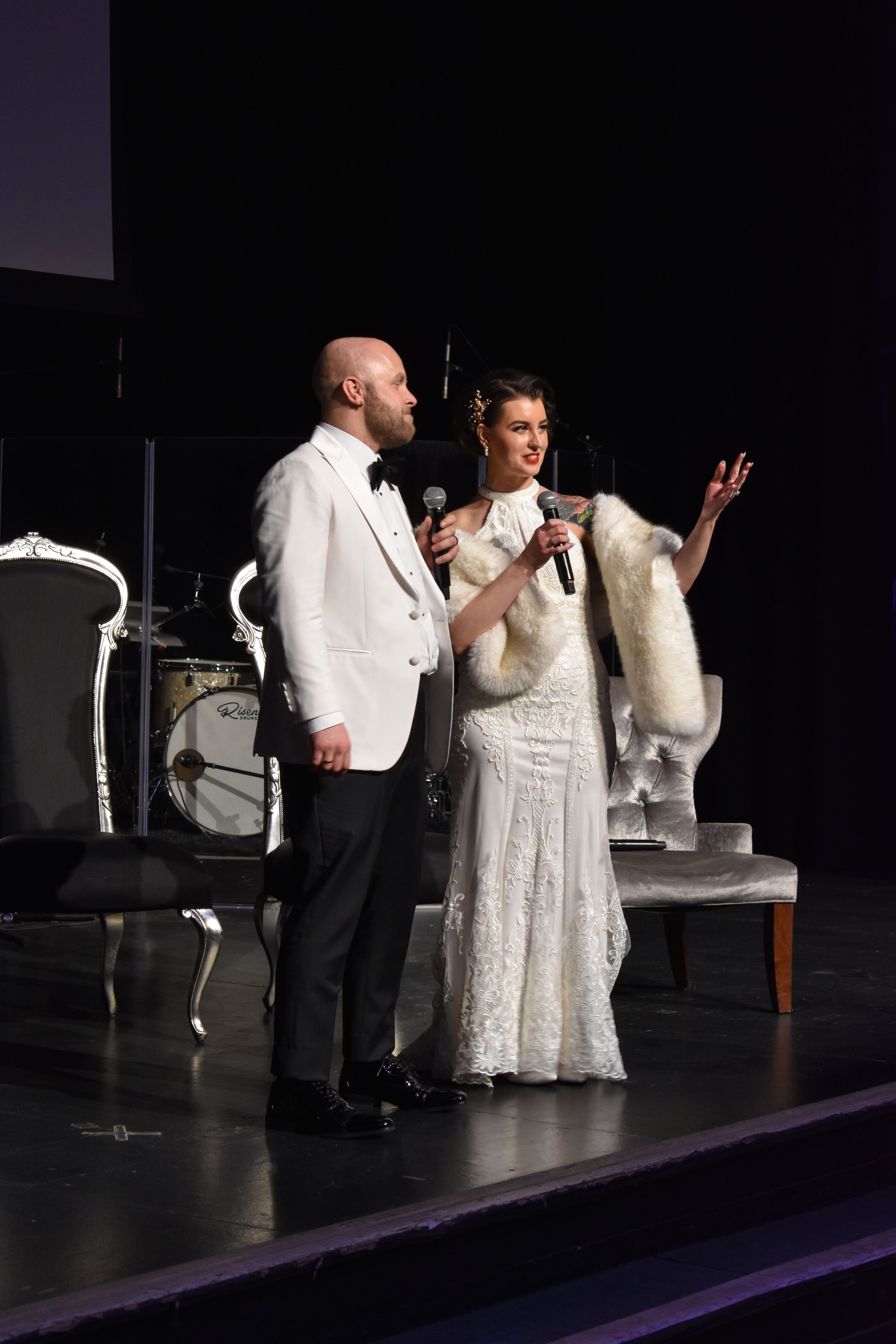 A man and woman dressed in formal wedding attire standing on stage with microphones, speaking or singing, in front of a dark background with a drum set and chairs.