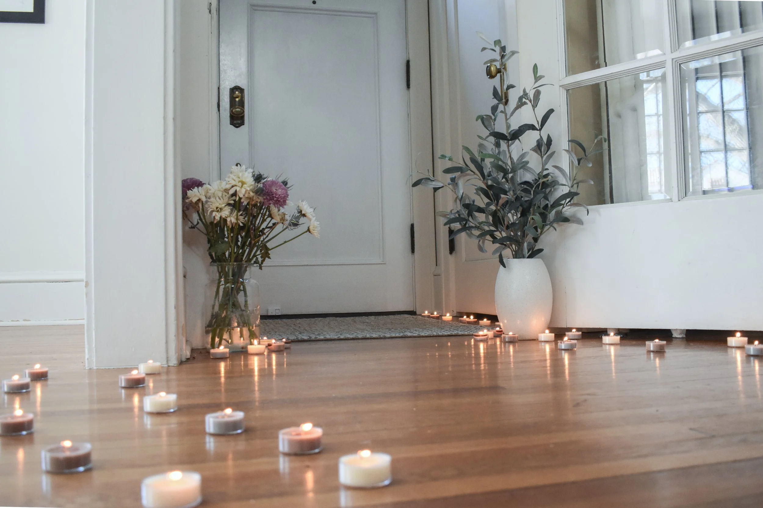 Decorative hallway entrance with candles on the wooden floor, a vase with flowers to the left, and a white vase with greenery to the right near a glass bay window.