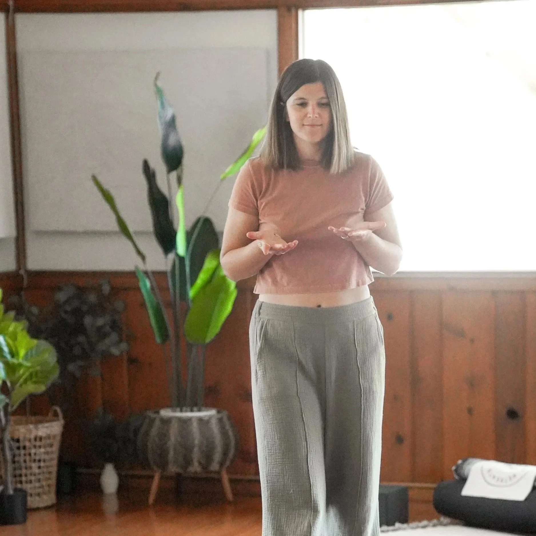 A woman with shoulder-length brown hair, wearing a cropped pink T-shirt and light-colored loose pants, standing indoors near a large window with sunlight streaming in, with potted plants and a whiteboard in the background.