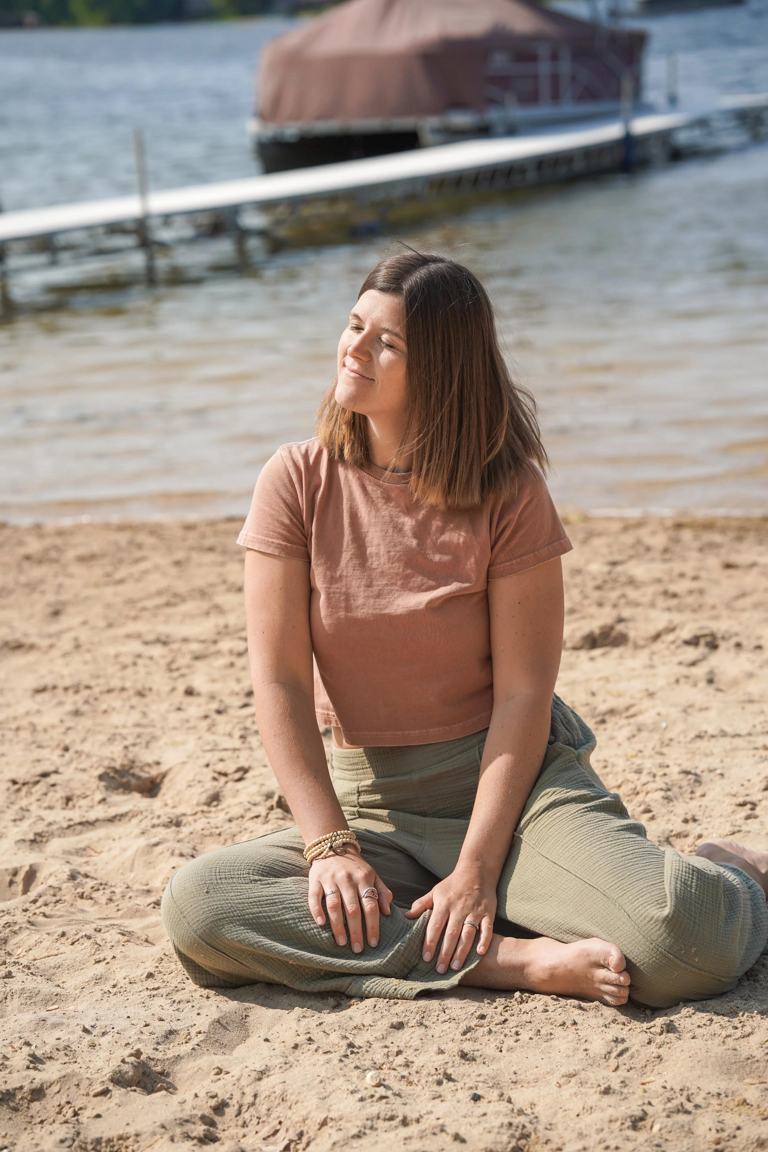 A woman sitting cross-legged on a sandy beach by the water with her eyes closed, enjoying the sunlight, with boats and a dock in the background.