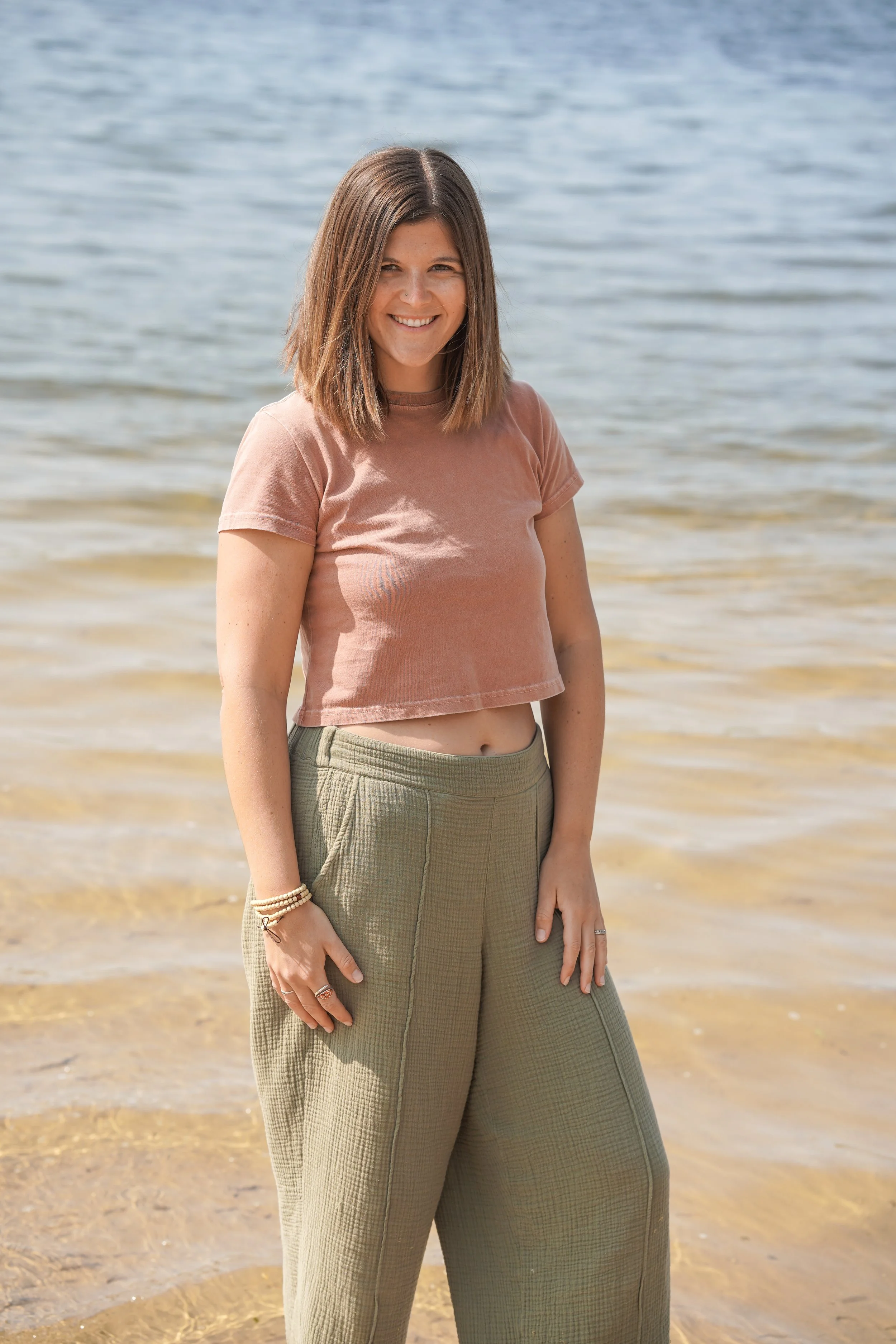 A young woman with shoulder-length brown hair smiling at the camera while standing on a sandy beach near the water.