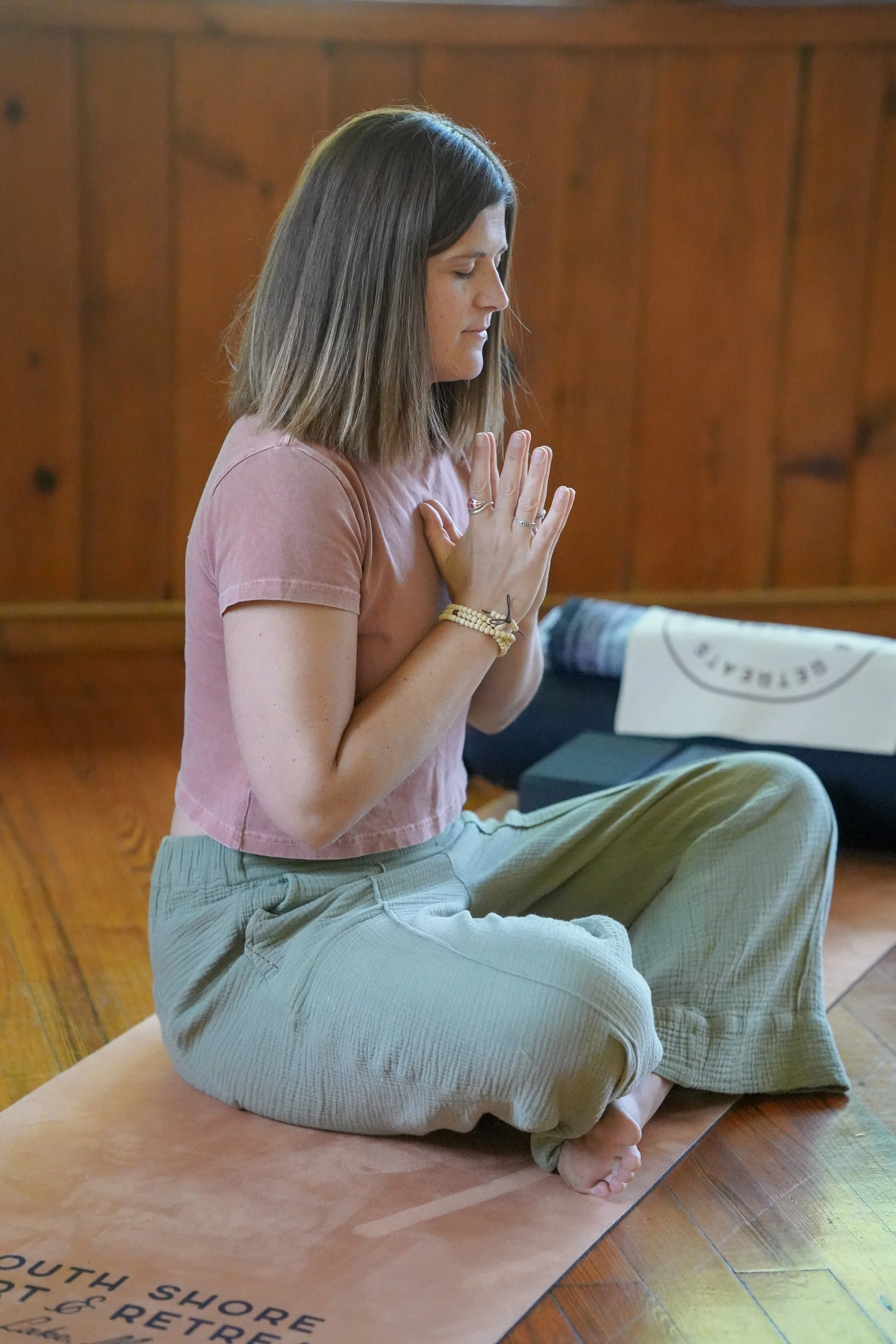 A woman sitting cross-legged on a yoga mat indoors, practicing meditation with her hands in prayer position, eyes closed, wearing a pink shirt and loose green pants.