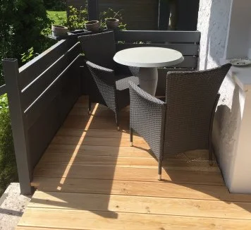 Ein Balkon mit Holzdecke, schwarzer Holzverkleidung, einem runden weißen Tisch und zwei schwarzen Korbstühlen, in der Nähe eine weiße Wand und grüne Bepflanzung im Hintergrund.