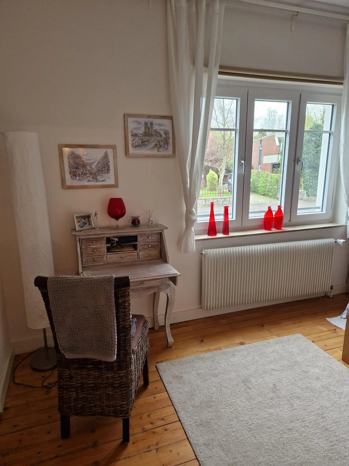 Ein Wohnzimmer mit Holzfußboden, einem weißen Teppich, einem Schreibtisch, einem Korbstuhl, roten Dekogläsern am Fenster, einer Stehlampe und Bildern an der Wand.