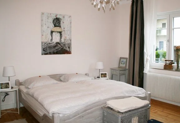 Schlafzimmer mit weißem Bett, Nachtschränken mit Lampen, Wandbild, Fenster mit Vorhängen, Heizkörper und Deko im Raum.