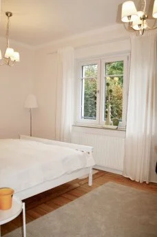 Ein Schlafzimmer mit weißem Bett, Fenster mit Blick auf Bäume, Lampe, Teppich und Vorhängen.