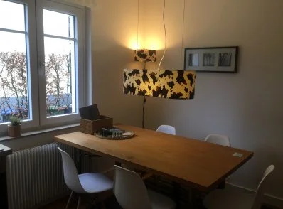 Ein Esszimmer mit einem Holztisch, vier weißen Stühlen, einem Fenster mit Blick nach draußen, einer Wandlampe und einer Wanddekoration mit drei Bildern.