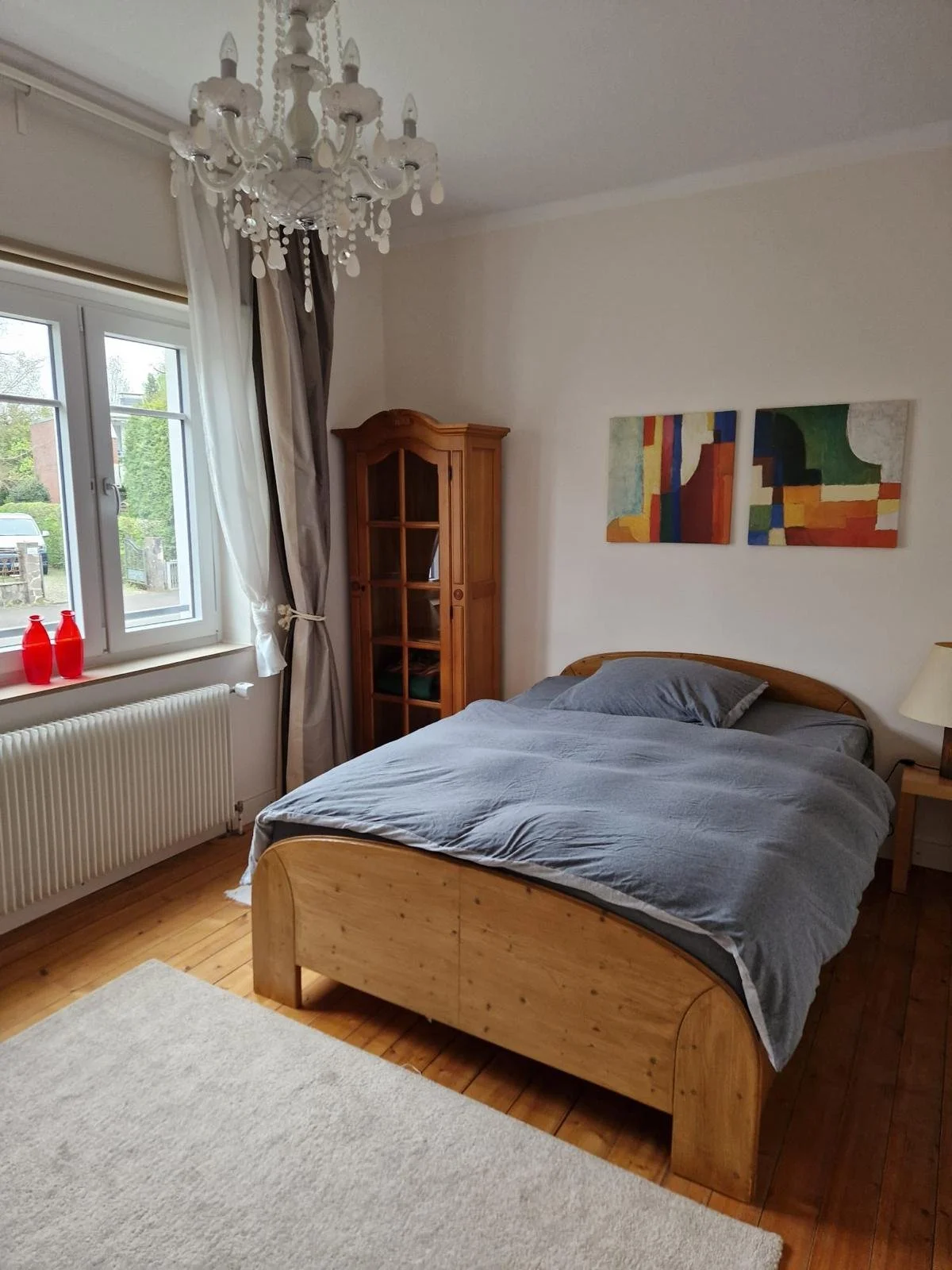 Schlafzimmer mit Holzbett, grauer Bettwäsche, Wandbilder, Holzschrank, Fenster mit Vorhängen, Kronleuchter, Teppich.