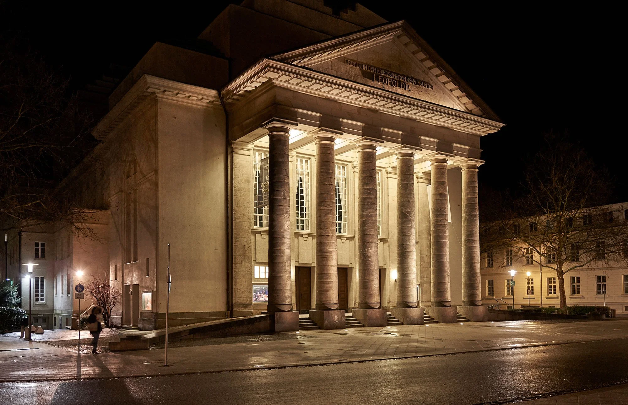 Ein illuminiertes neoklassizistisches Gebäude mit großen Säulen bei Nacht, umgeben von ruhiger Straße und Bäumen.