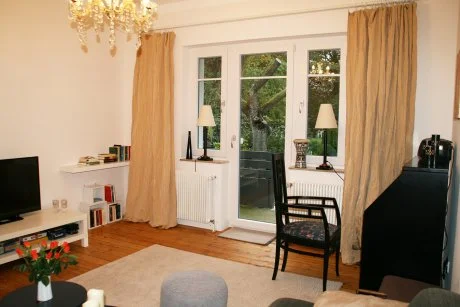 Wohnzimmer mit großen Fenstern, Gardinen, Sessel, Fernseher, Kirschblumen und einem Blick nach draußen auf Bäume.