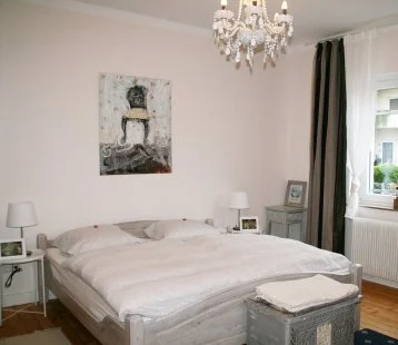 Schlafzimmer mit weißem Bett, Nachttischen, Lampen, Bild an der Wand, Fenster mit Vorhängen und Dekorationen.