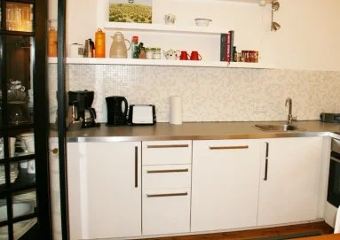 Küchenzeile mit weißen Schränken, schwarzen Küchenutensilien, Toaster, Kaffeemaschine, Geschirrspüler, Spüle und Regal mit verschiedenen Gegenständen