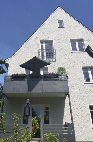 Mehrstöckiges weißes Haus mit Balkon, Dach und Garten, blauer Himmel.