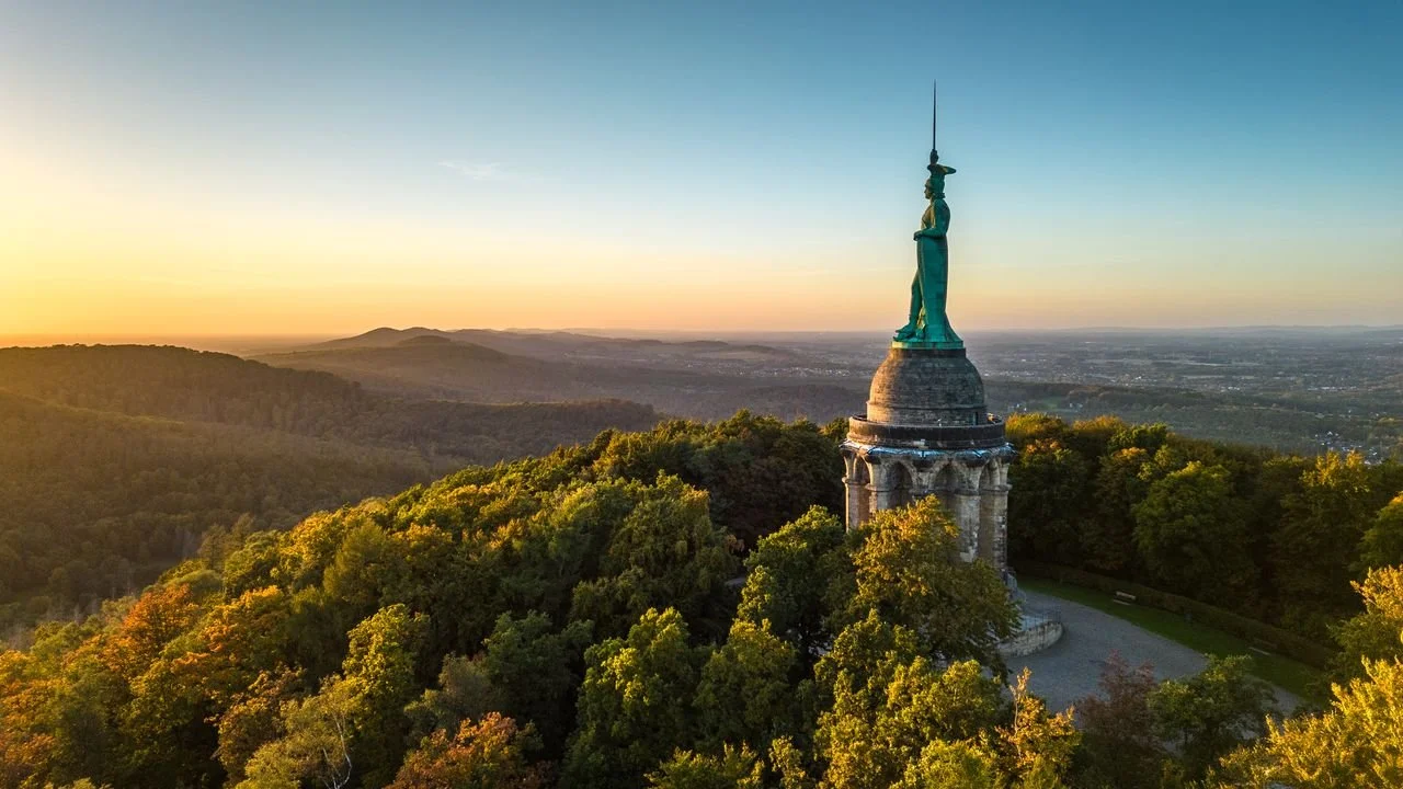 Fotografische Aufnahme einer Statue auf einem Hügel bei Sonnenuntergang, umgeben von Bäumen, mit einer weiten Landschaft im Hintergrund.